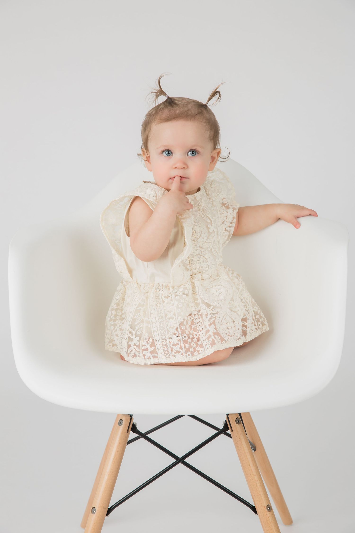 A toddler in a white lace dress sits on a modern white chair with finger near mouth.