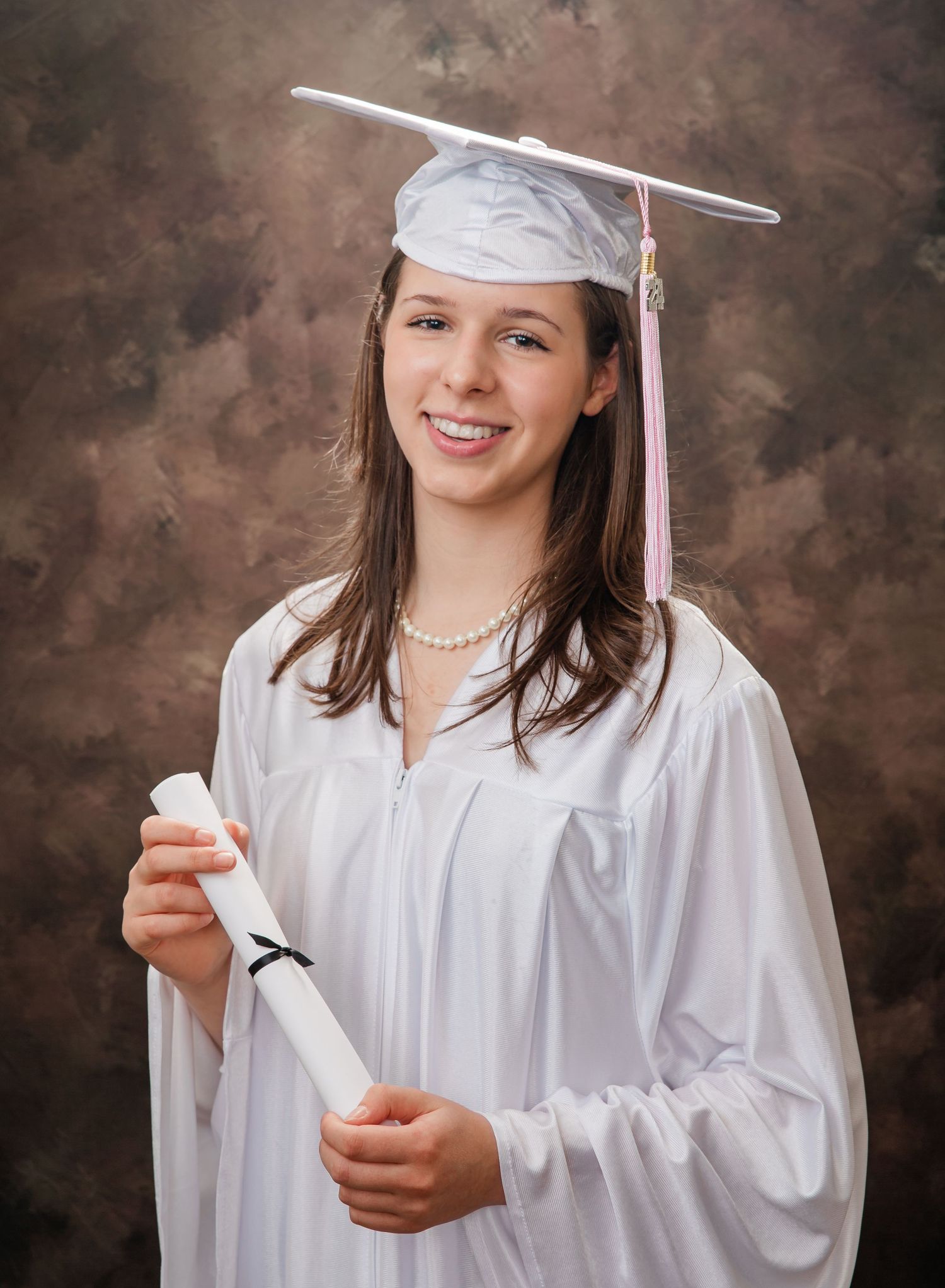 A graduate in white cap and gown holds a diploma while posing for a traditional portrait against a brown backdrop.