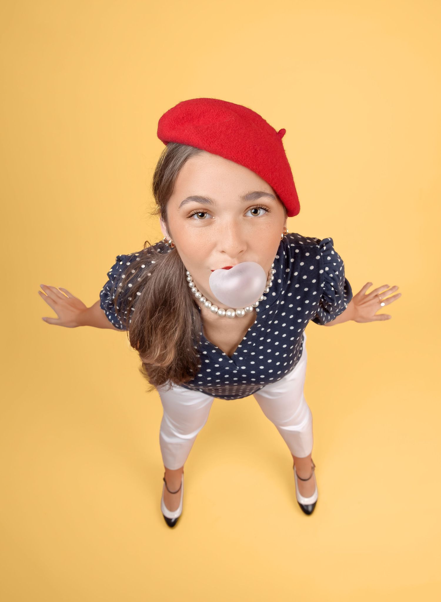 Fun wide-angle view of someone in a red beret and polka dot top with outstretched arms against a yellow backdrop.