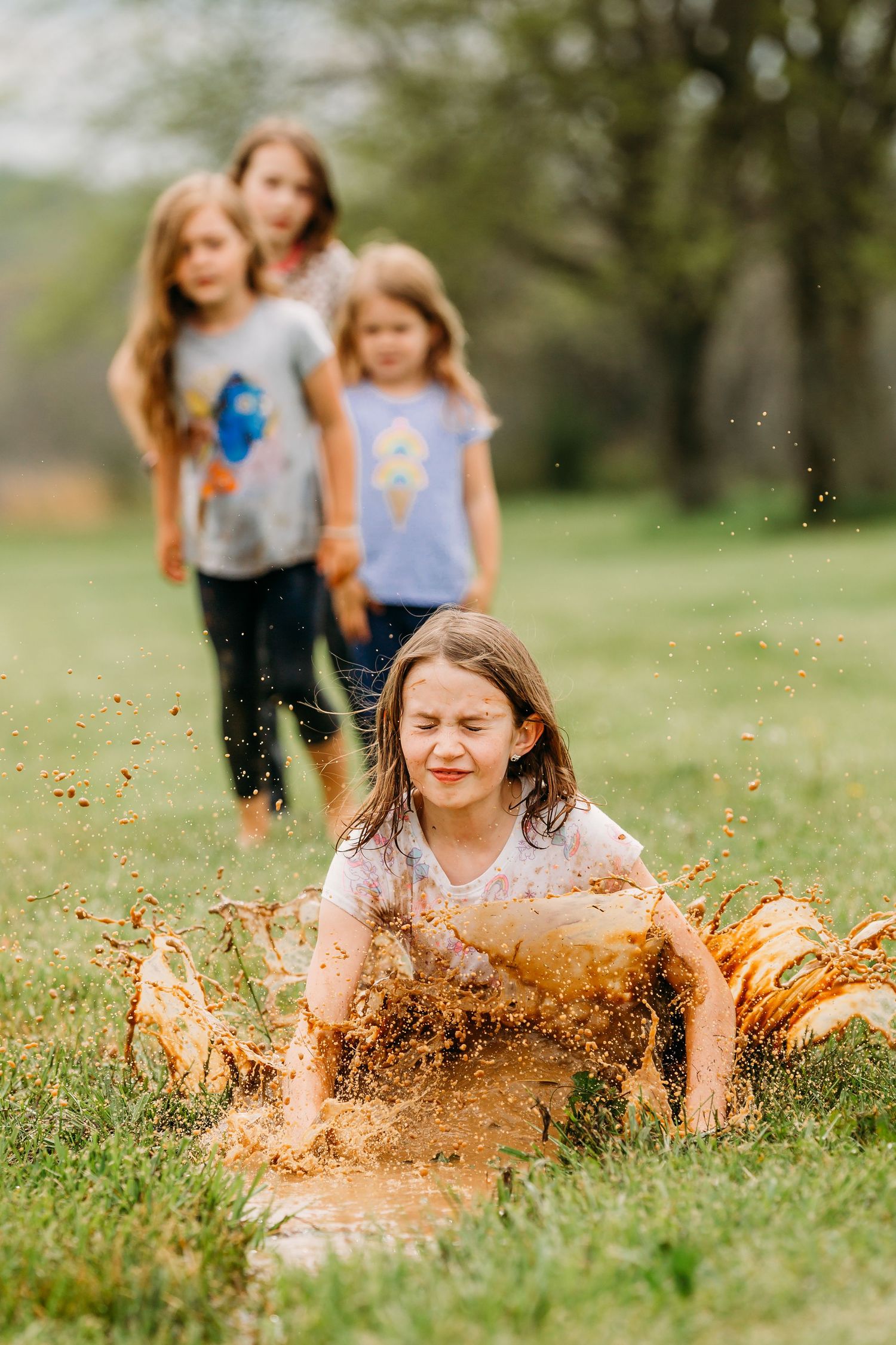 Children playing outdoors as someone splashes in a mud puddle creating a dramatic spray effect in a grassy field.