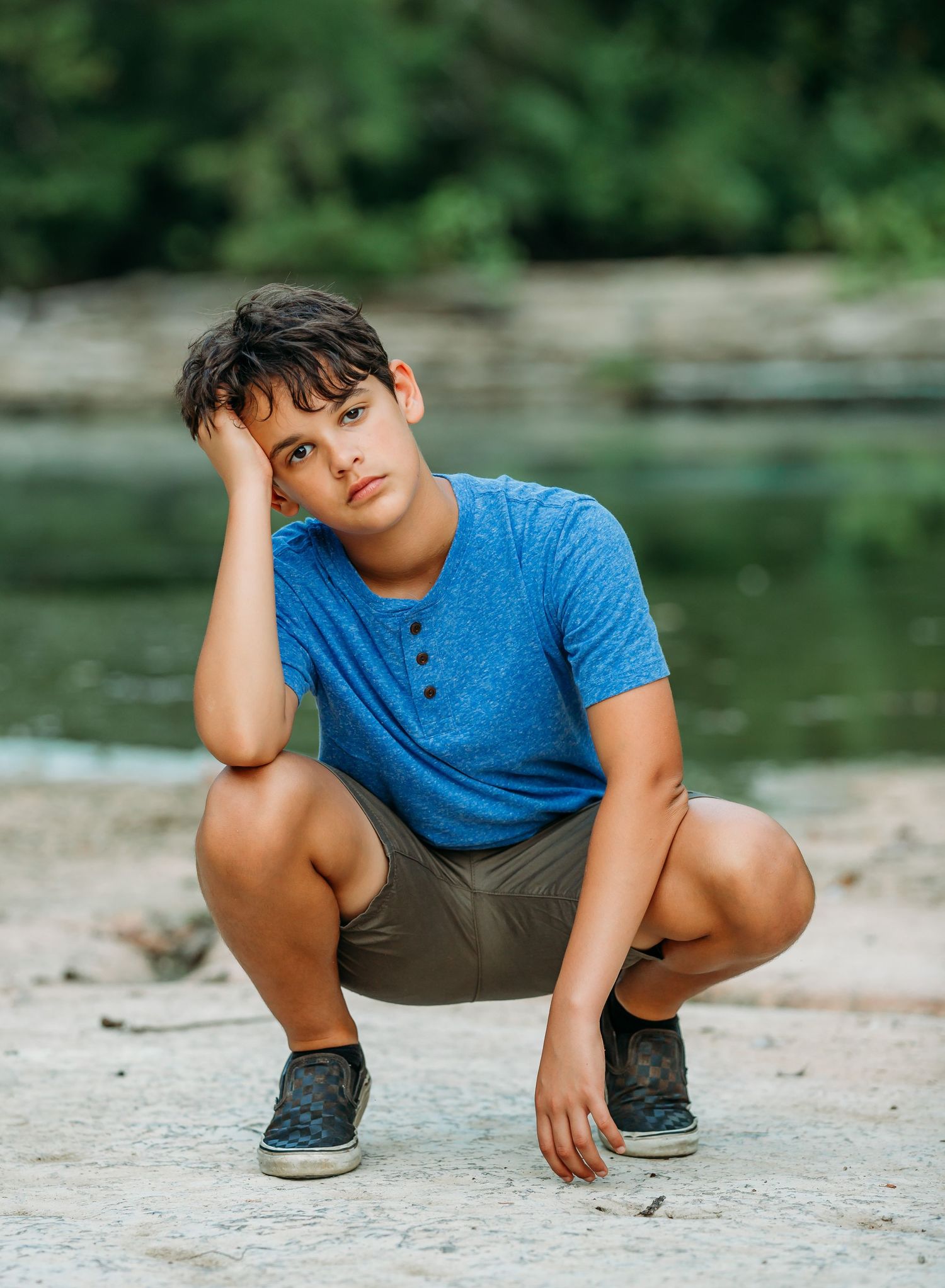 Young person in blue shirt crouches by waterfront with hand on head in casual pose.