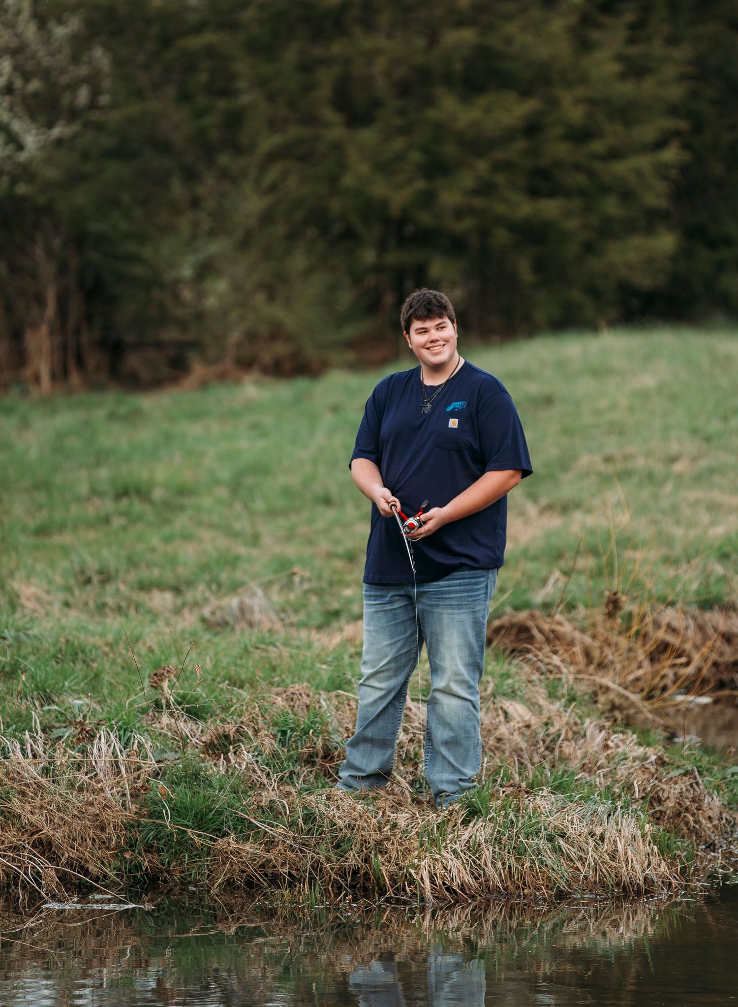 Person in navy shirt and jeans stands near water's edge in green grassy outdoor setting.