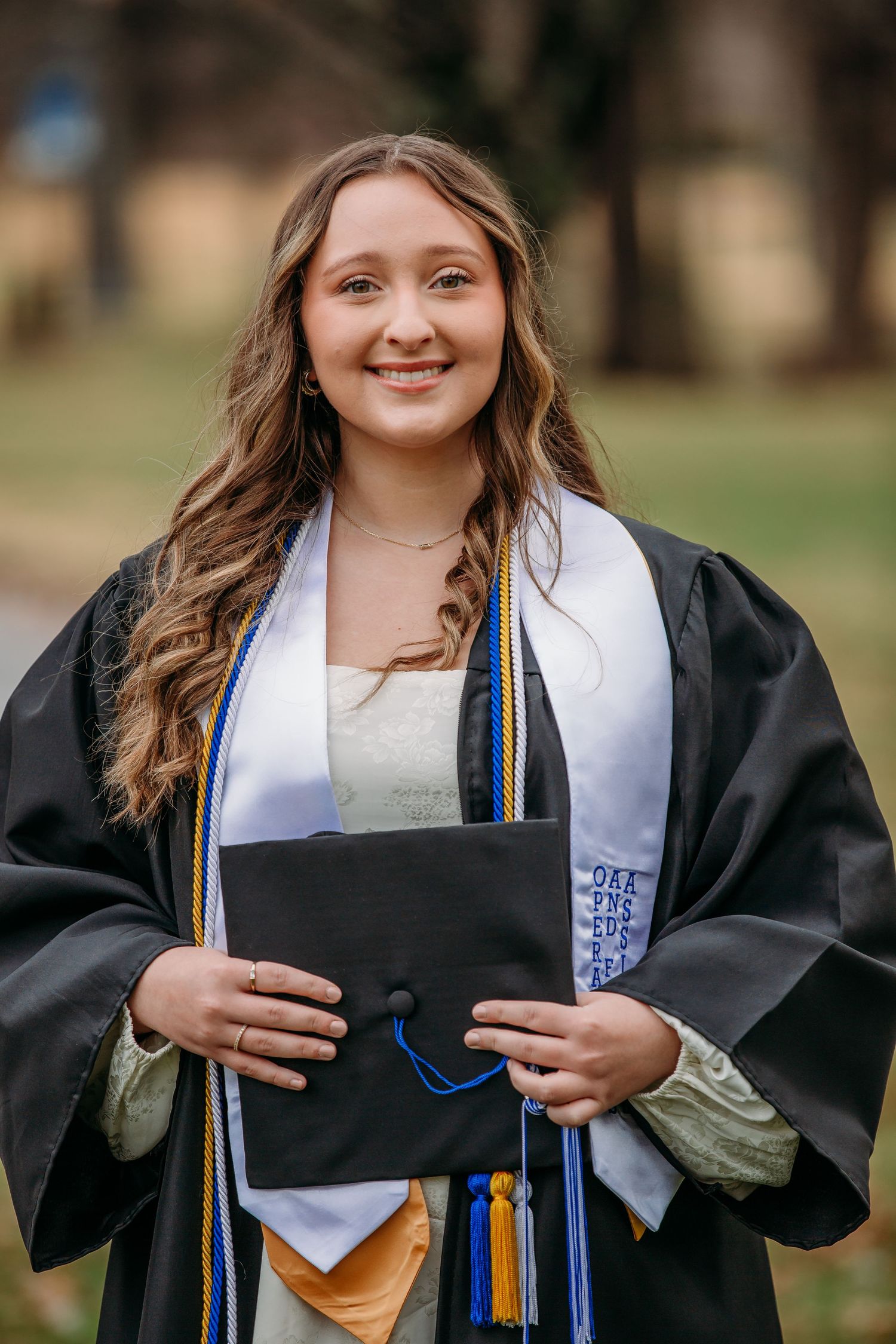 Graduate in traditional black cap and gown holds degree while smiling during outdoor graduation photo session.