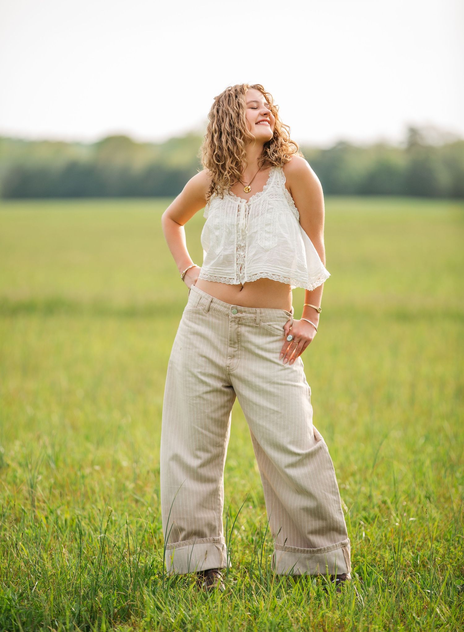 Person in white crop top and wide-leg khaki pants stands joyfully in a sunlit green meadow during golden hour.