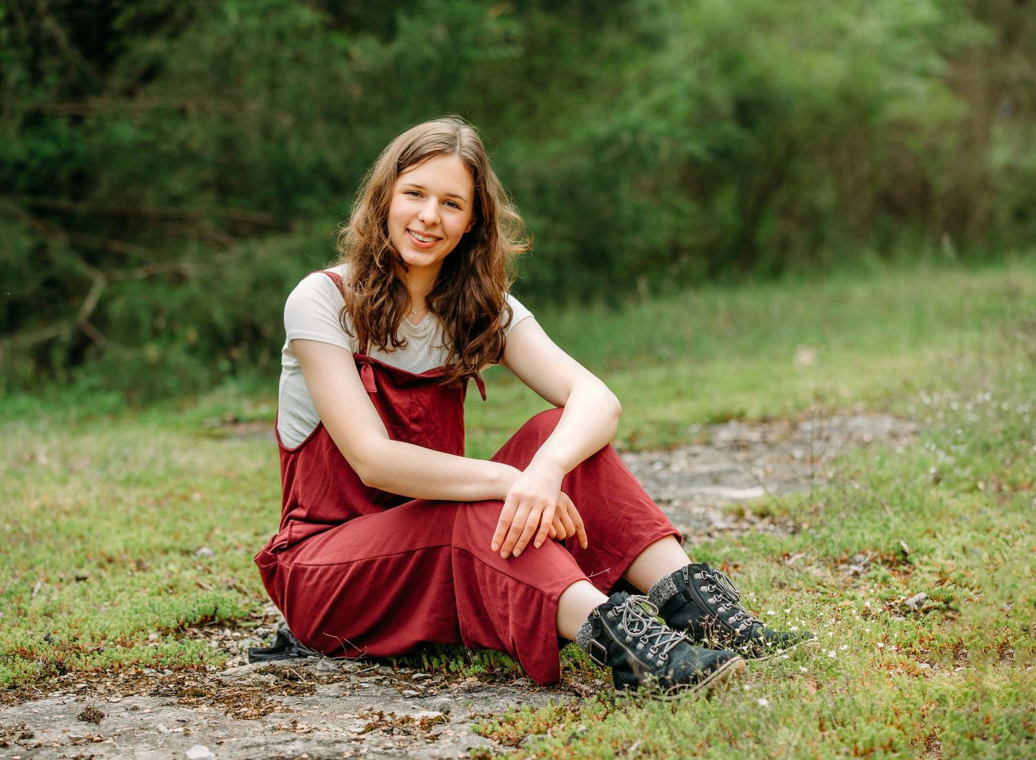 Person in a burgundy dress sits on mossy ground in a naturally lit outdoor setting during a photo session.