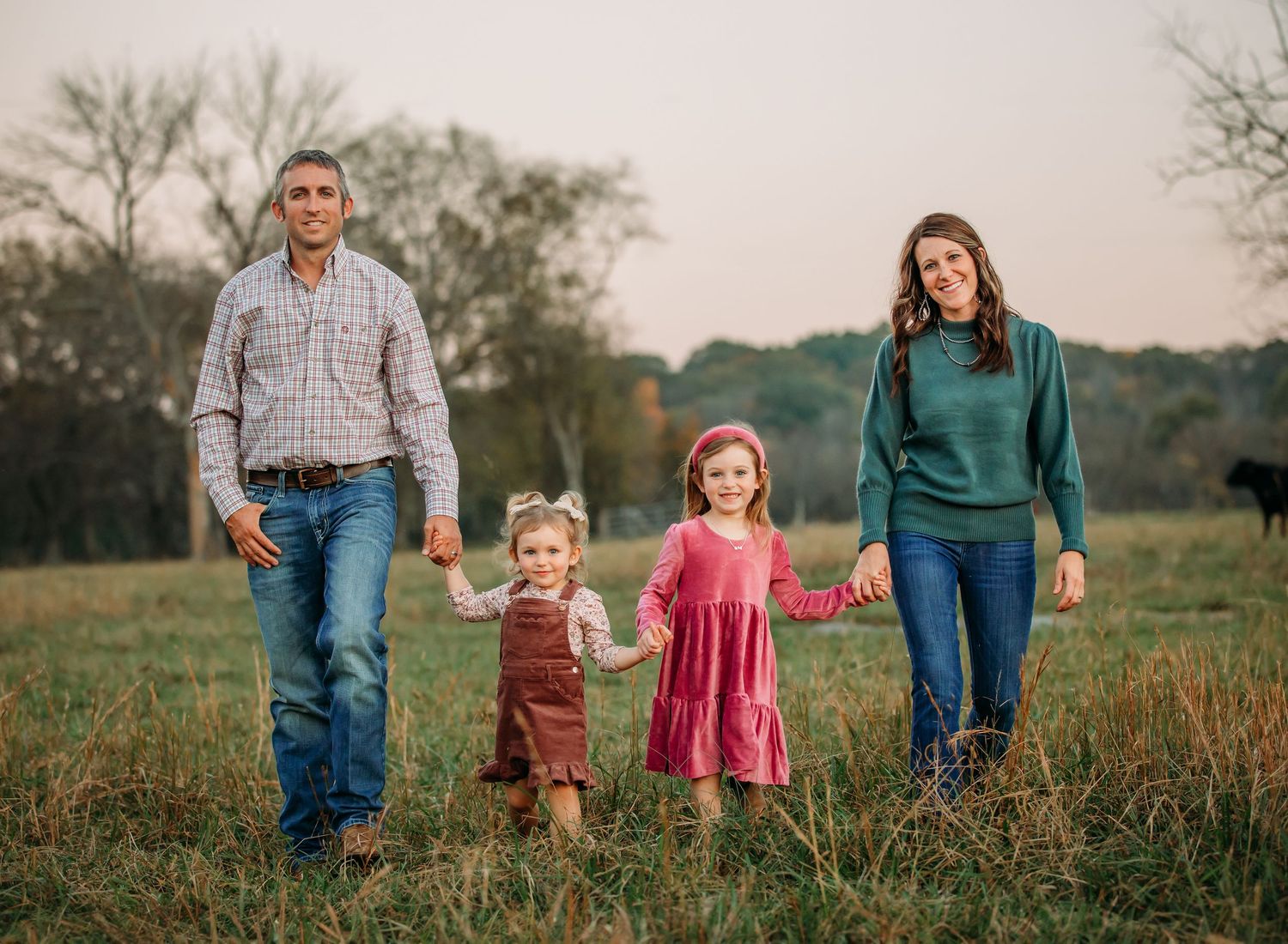 A family of four holds hands while walking together in a grassy field at sunset.