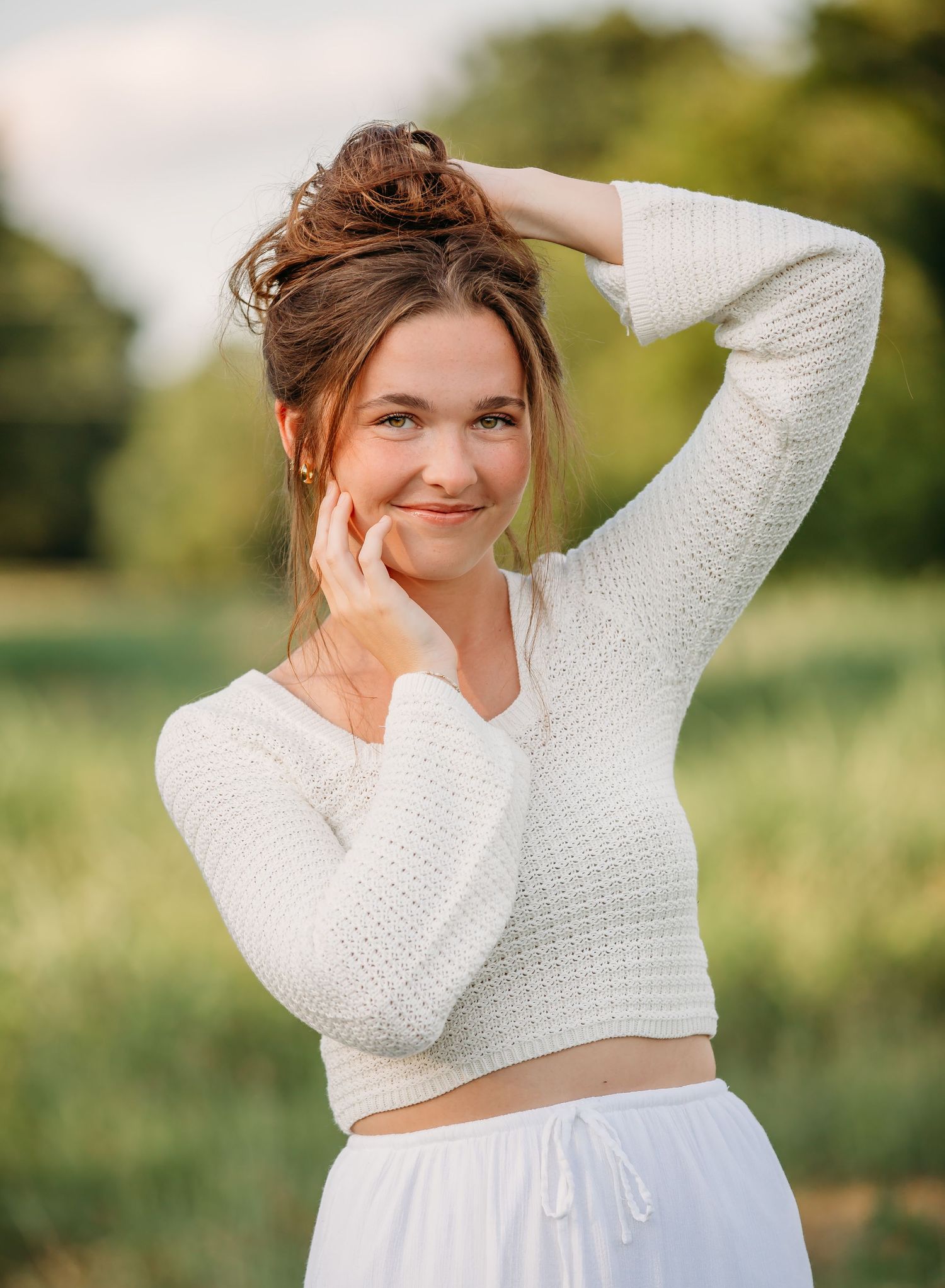 Someone in a white knit crop top and white skirt poses outdoors in a sunny field with natural lighting.