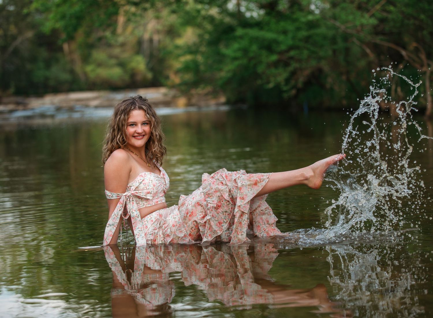 Person in floral dress sitting in lake water makes playful splash while posing during outdoor photo session.