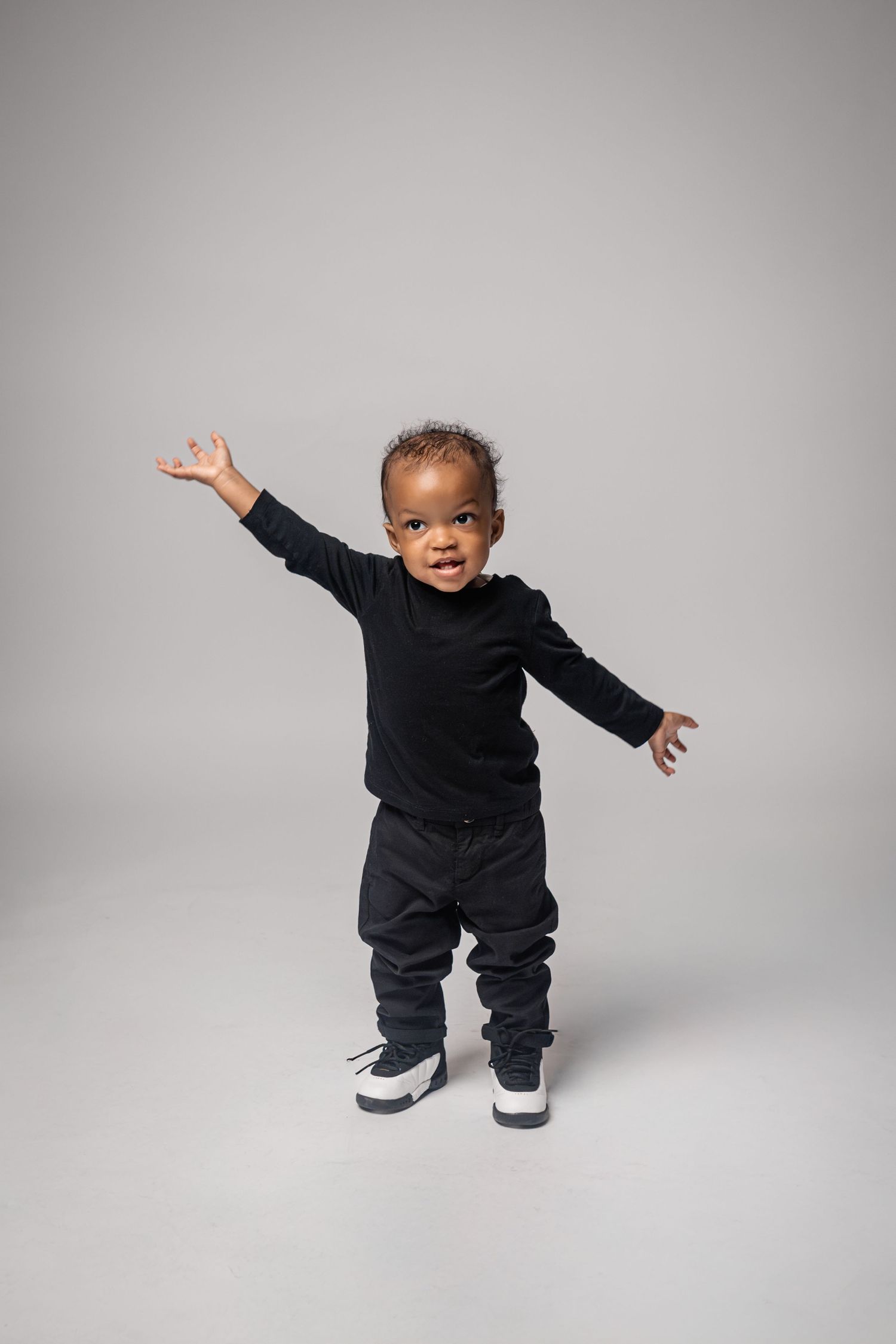 Baby in black outfit learning to walk and dance against gray backdrop in studio setting.