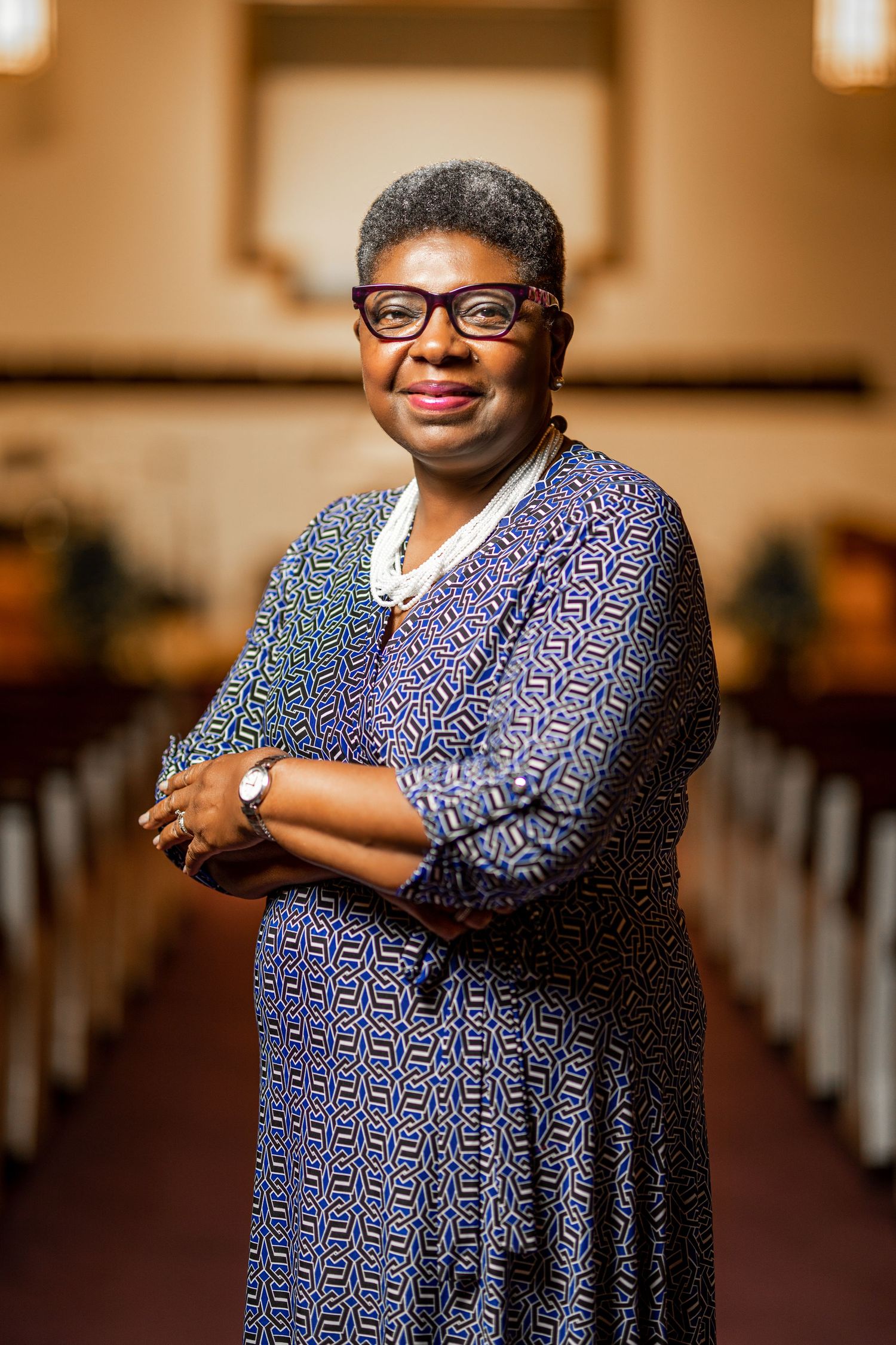 A woman wearing a blue and purple patterned dress and glasses poses confidently in a warm-lit church setting.