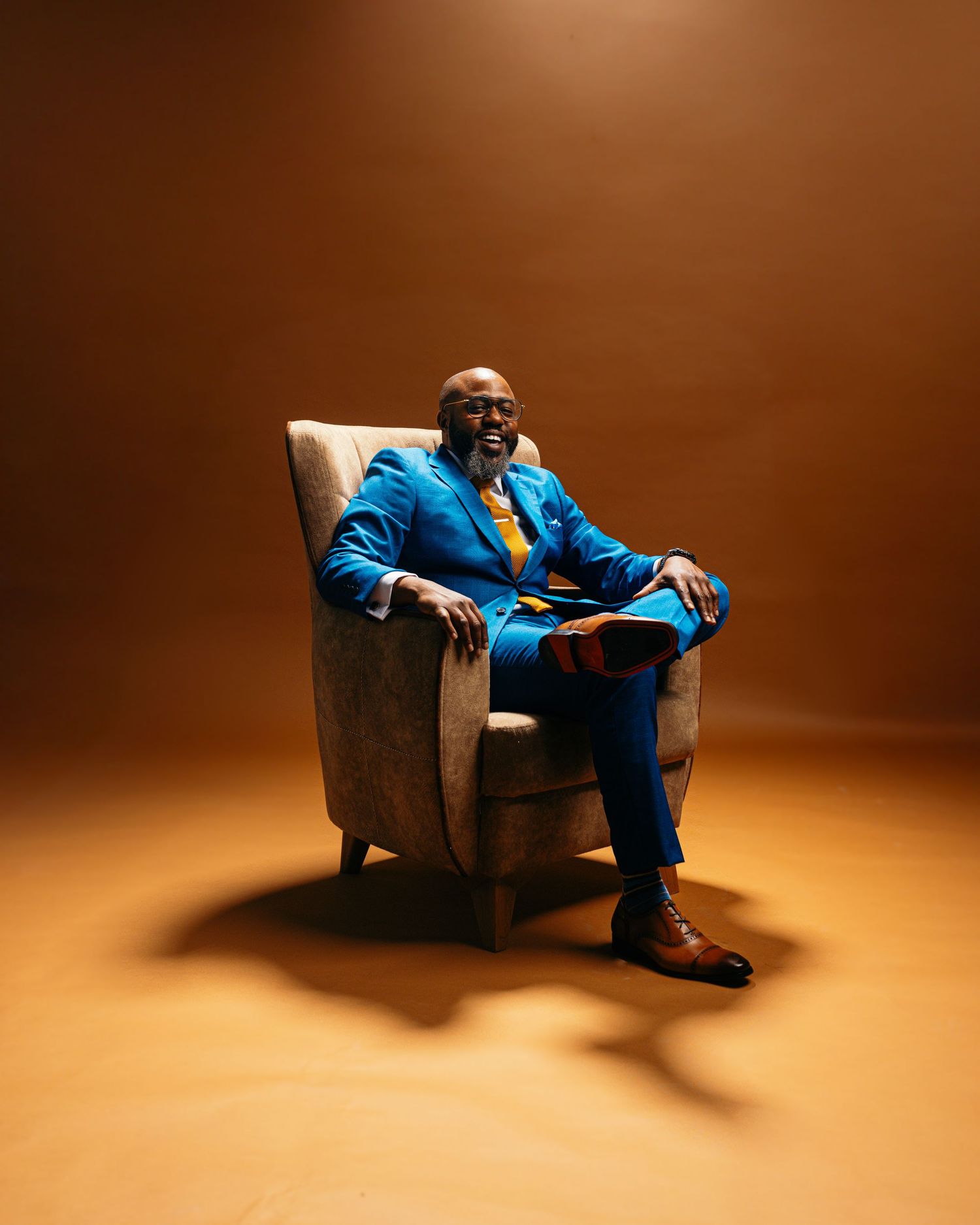 Studio photos of someone in a bright blue suit lounging in an armchair against a warm brown backdrop.