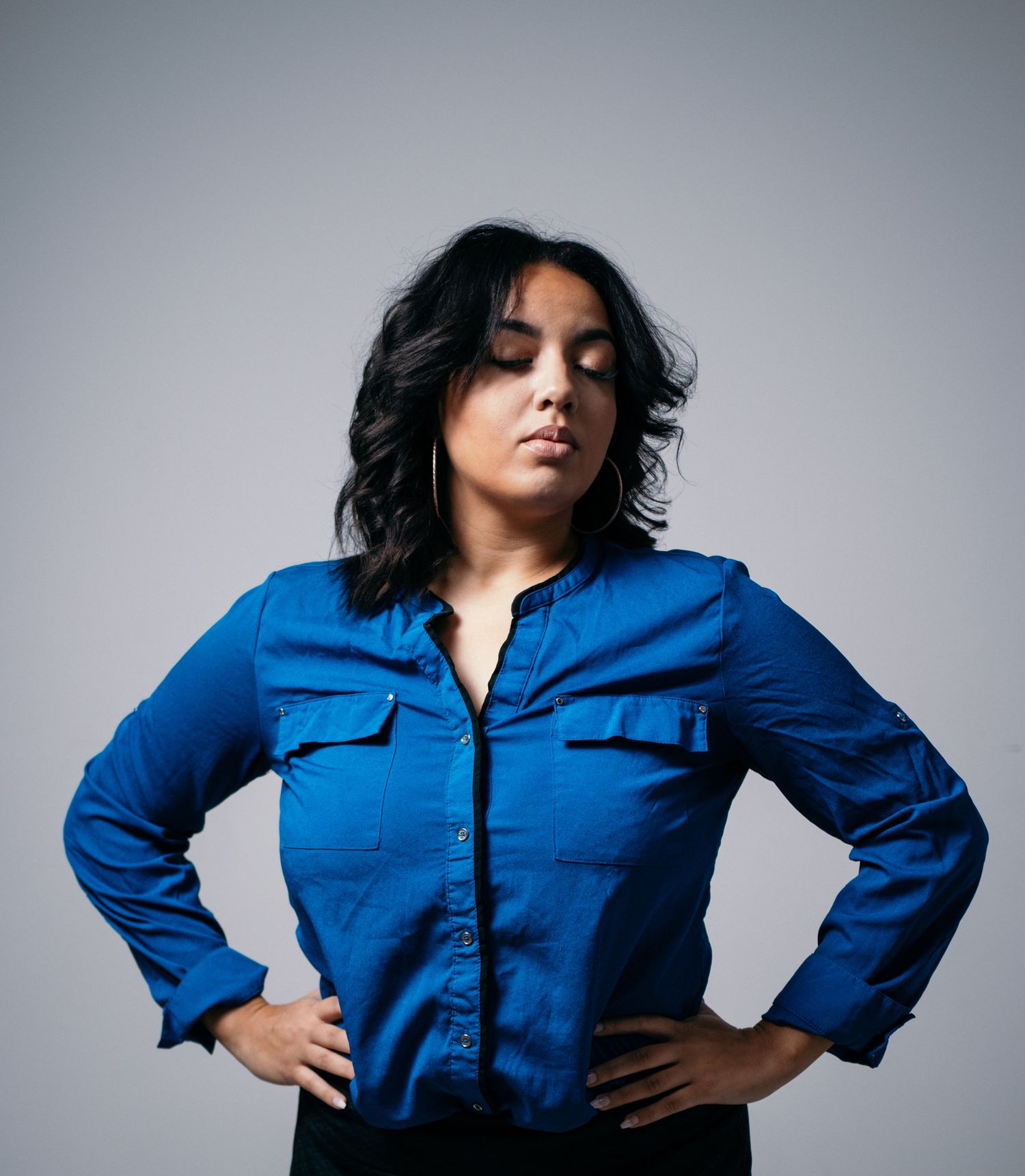 Woman in royal blue button-up shirt poses with hands on hips against grey background.