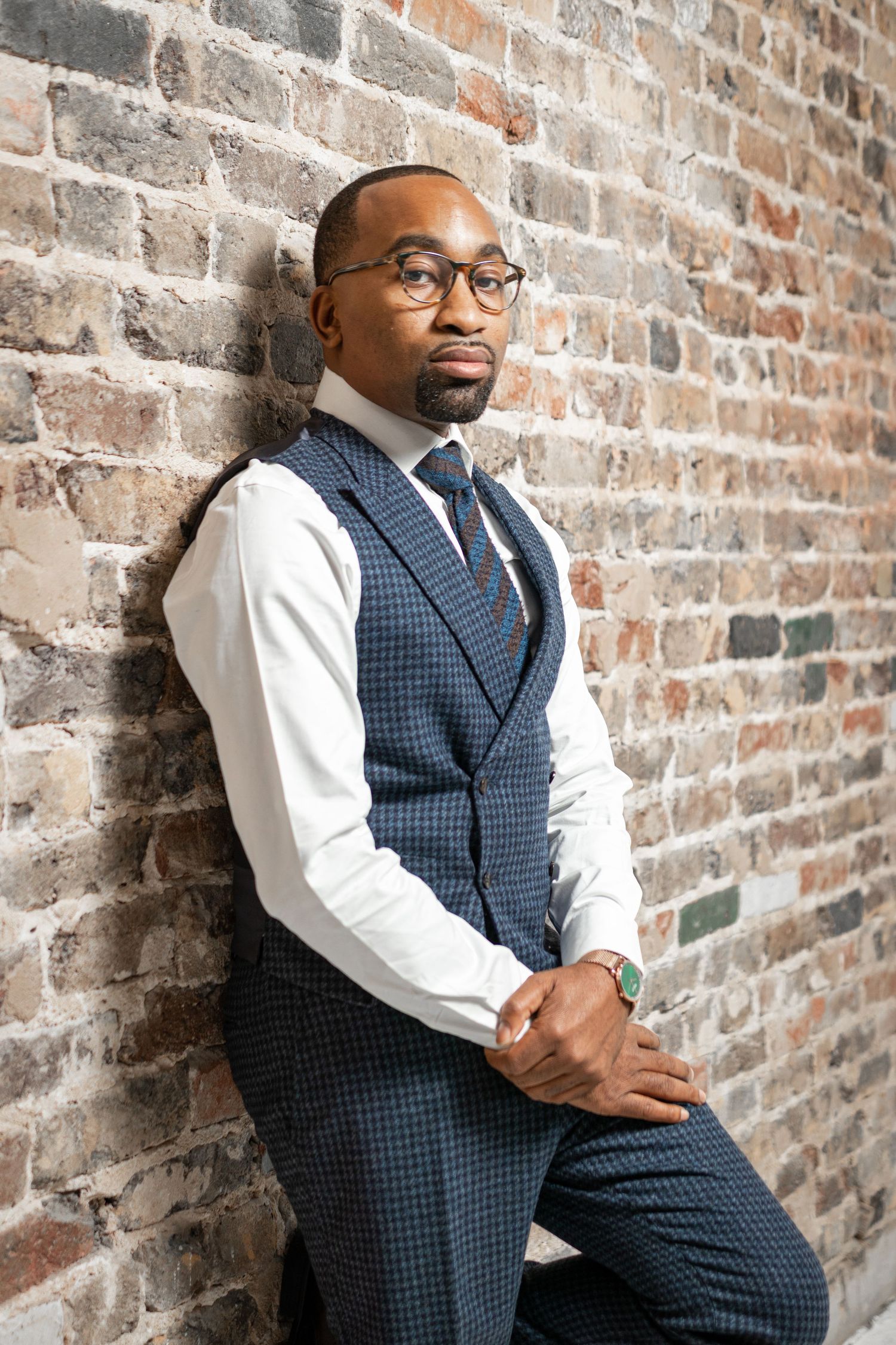 Professional man in blue checkered vest and white shirt poses against brick wall.