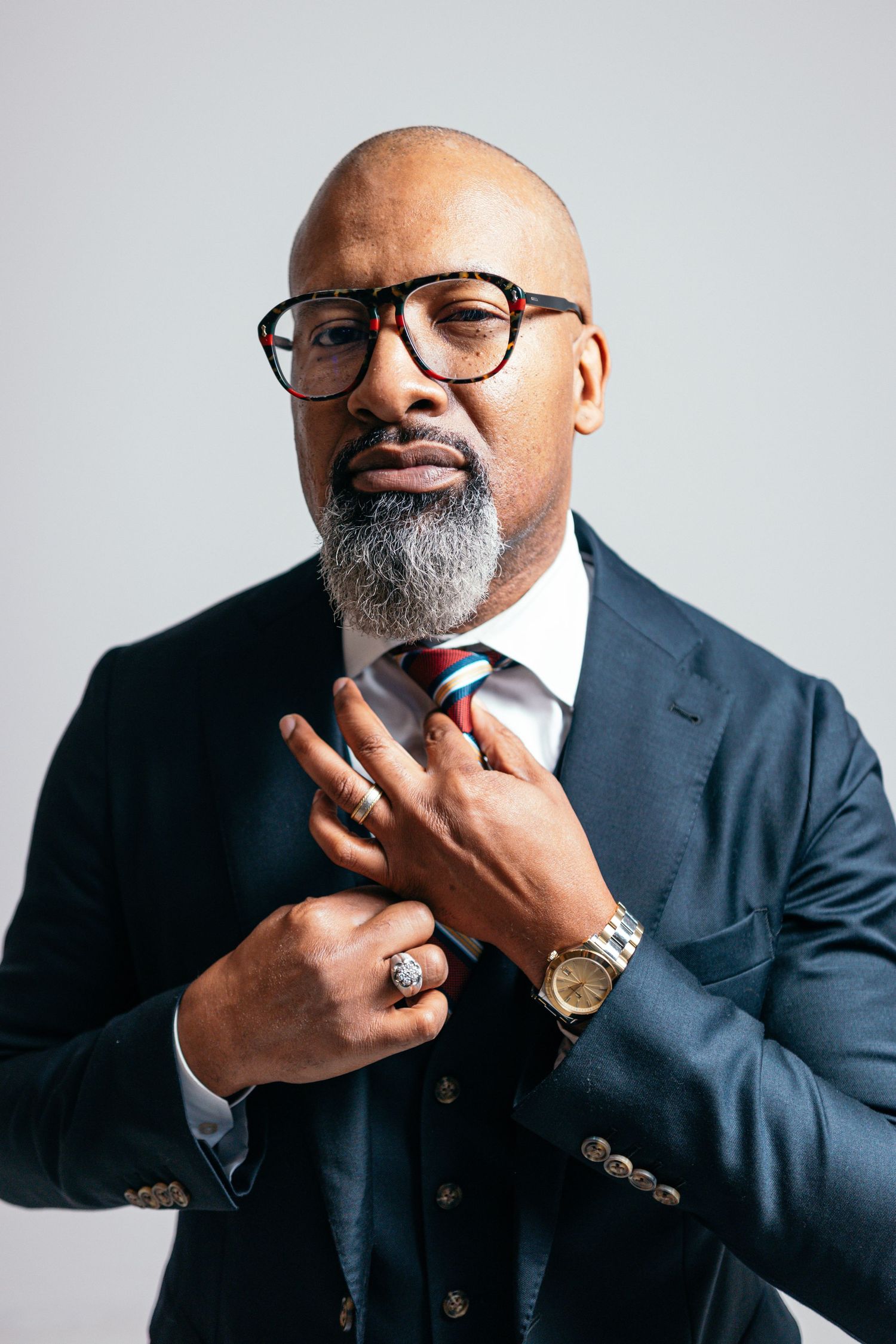 Distinguished man in suit and glasses touches tie while posing against grey background.