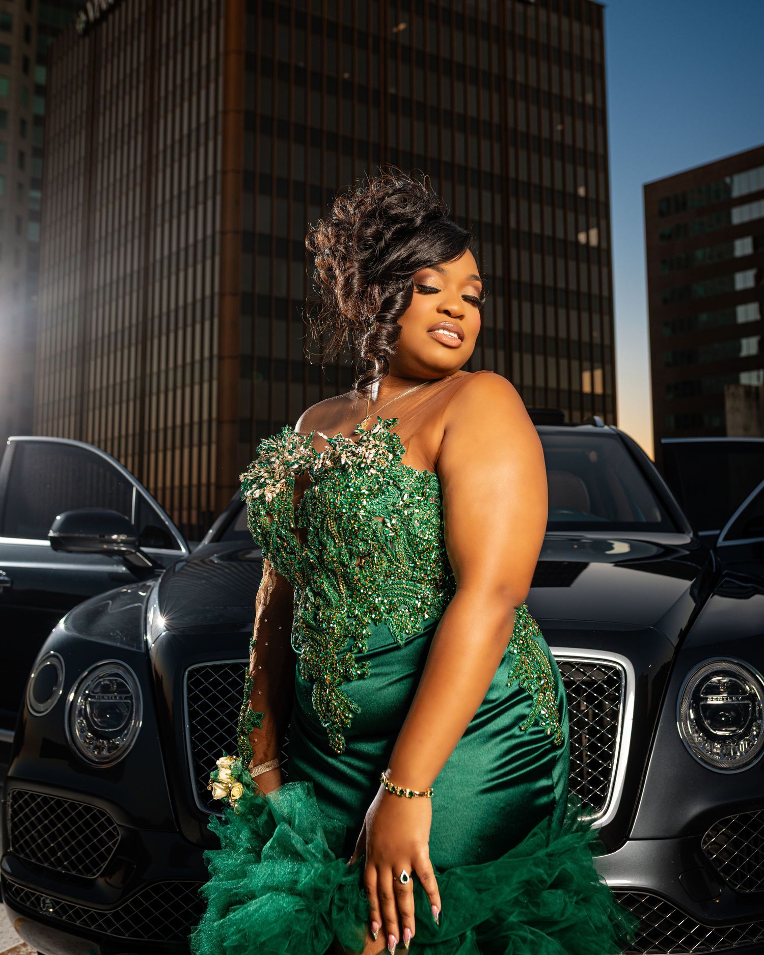 Woman in sparkly emerald green evening gown poses dramatically next to luxury cars.