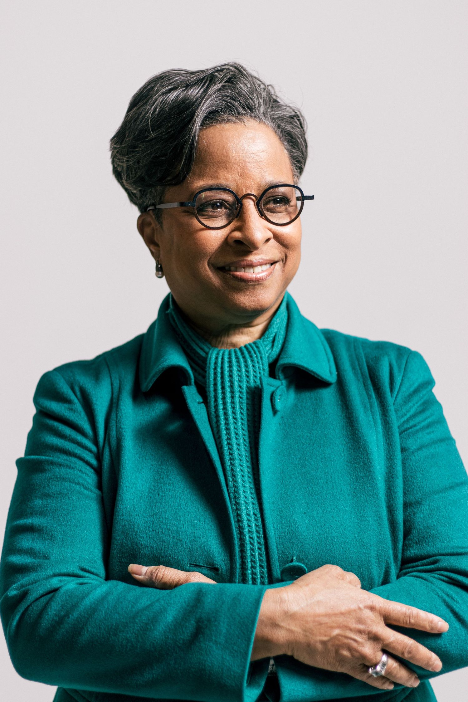 Professional in teal jacket and glasses smiling warmly against light background.