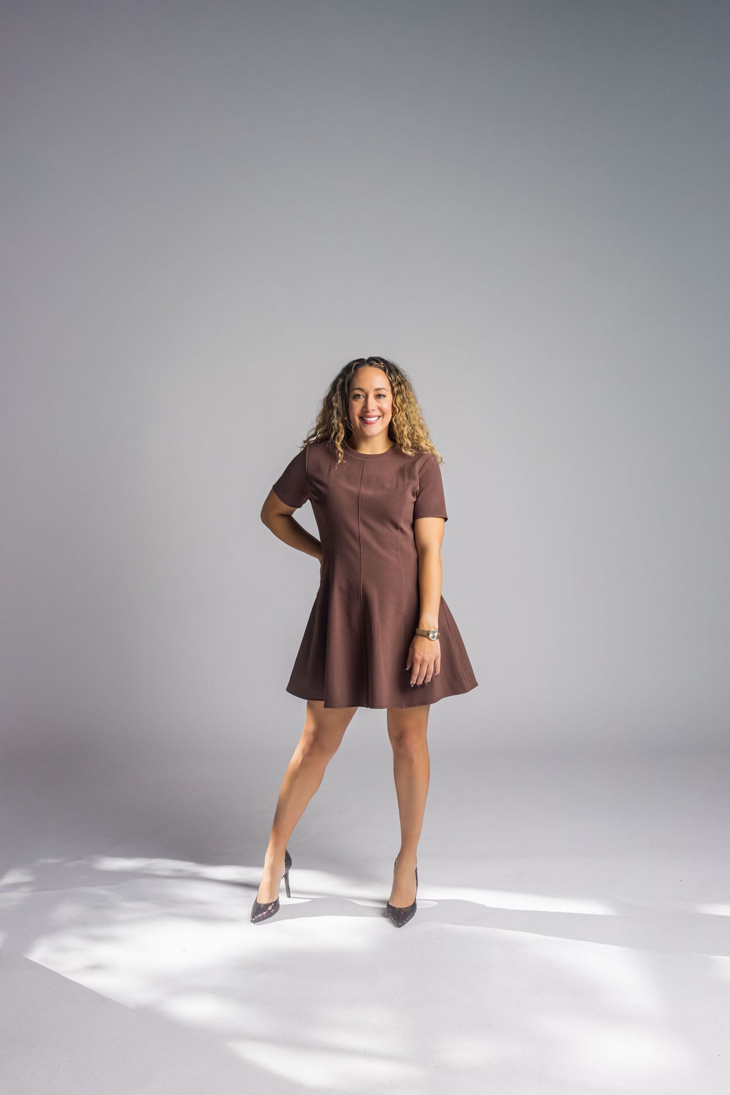 Stylish woman in brown dress poses confidently on grey background.