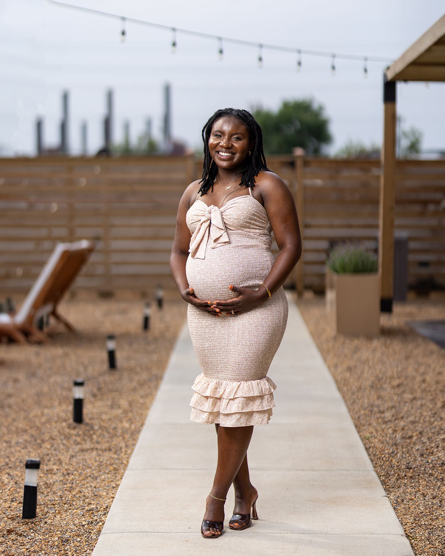 Fashion photography on a wooden deck wearing a pink ruched dress and string lights overhead.
