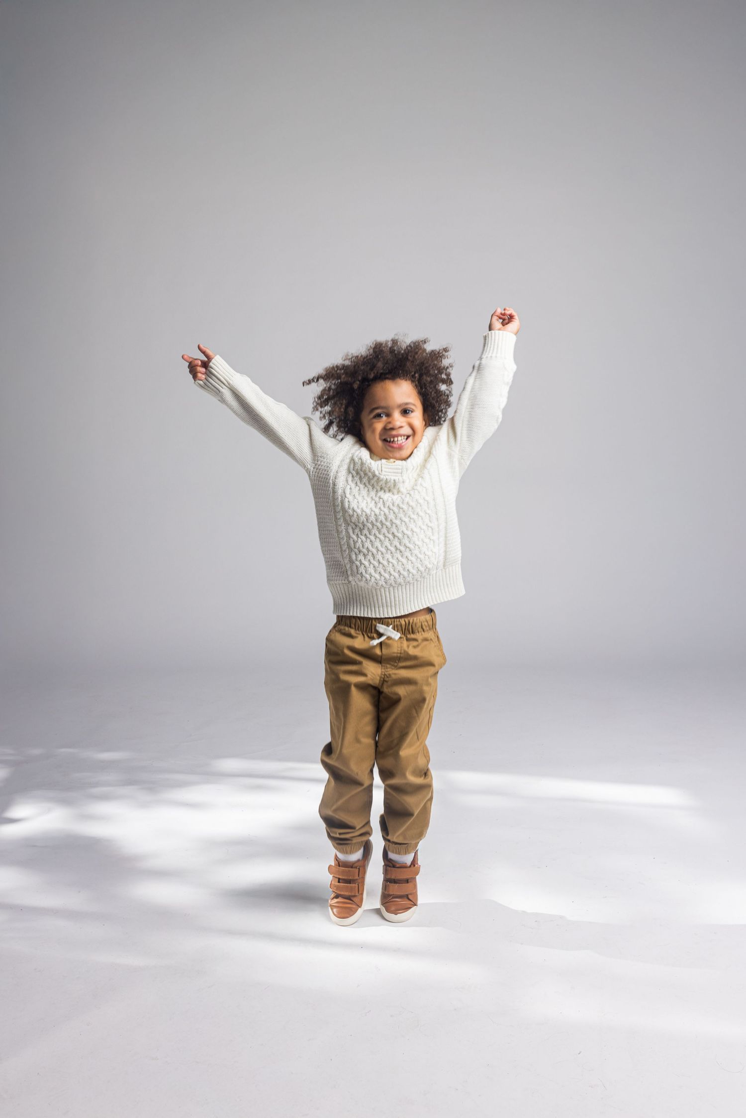 Energetic child in white sweater and brown pants jumping joyfully against grey background.