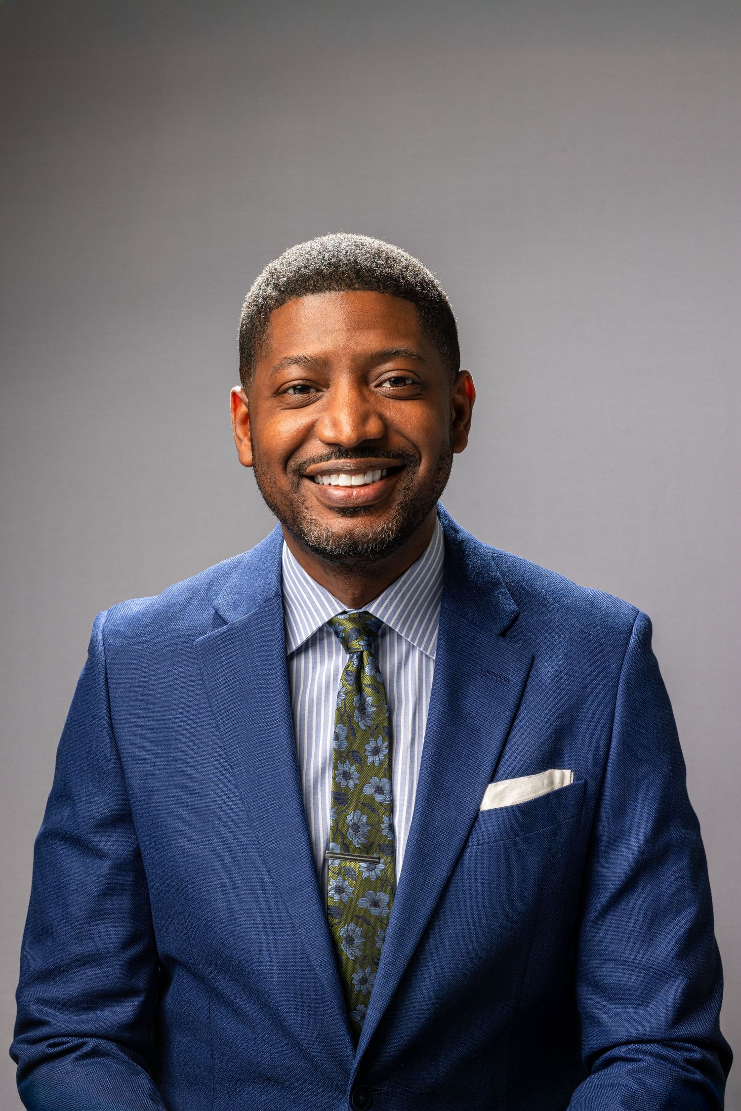 Professional man in blue suit and patterned tie smiling warmly against grey backdrop.