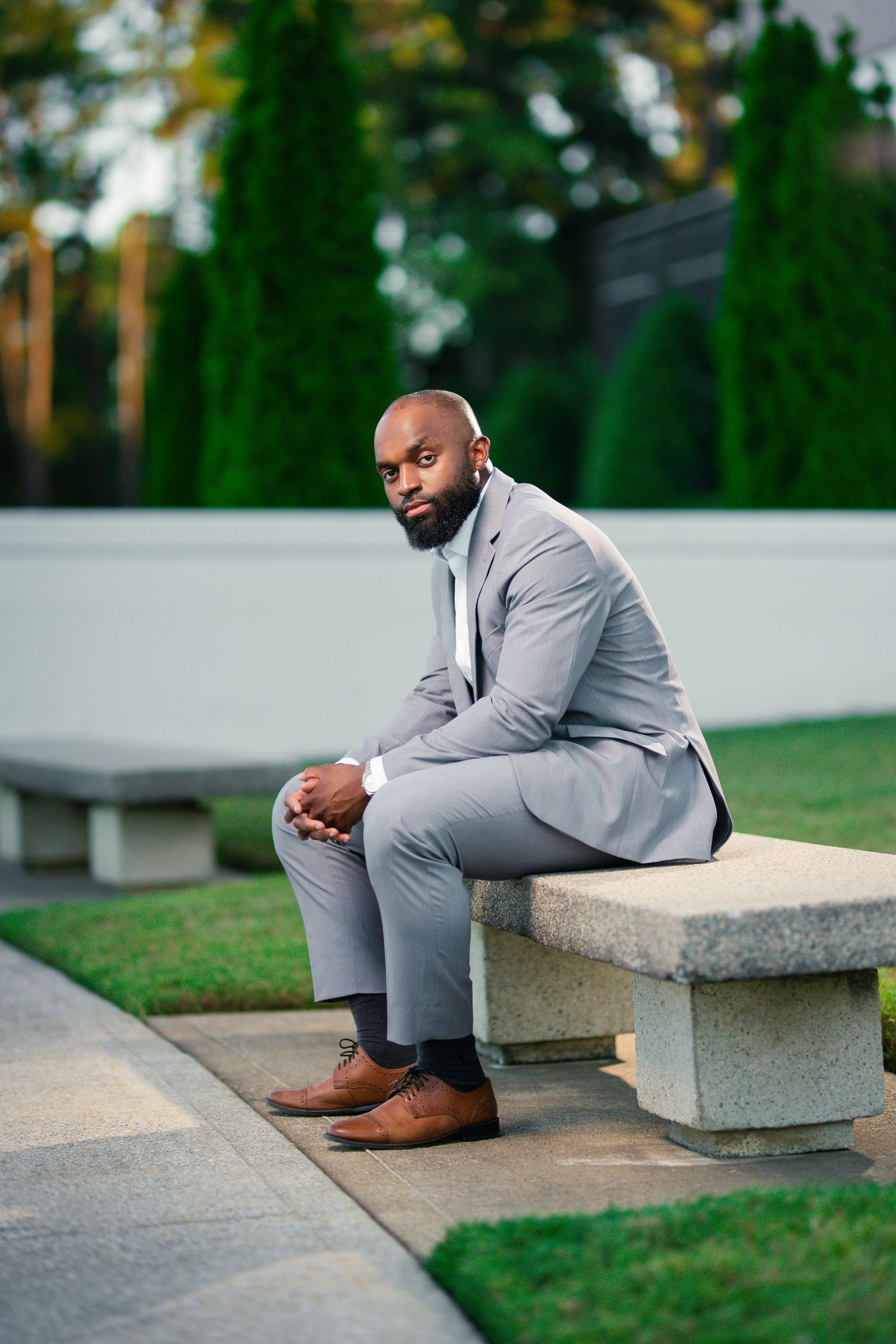 Professional portrait in light grey suit sitting on concrete bench in garden setting.