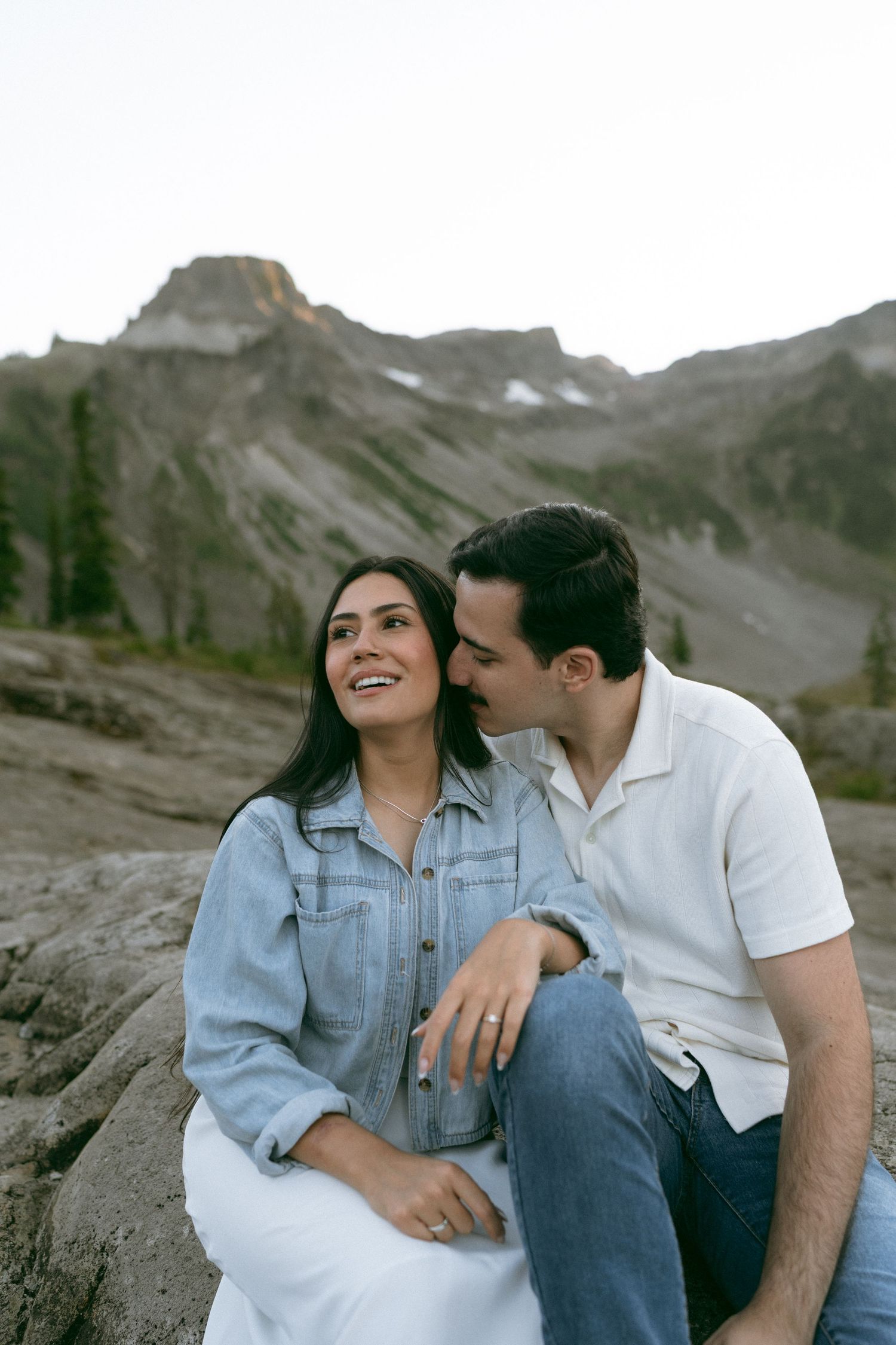 Smiling couple sits close on a rocky mountainside as a man kisses the cheek of a woman in a denim jacket, with rugged peaks behind them.