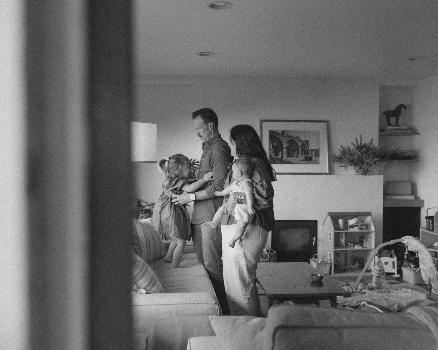 Black and white photo of a family of four gathered in a cozy living room with a sofa, framed art, shelves, and a small television.