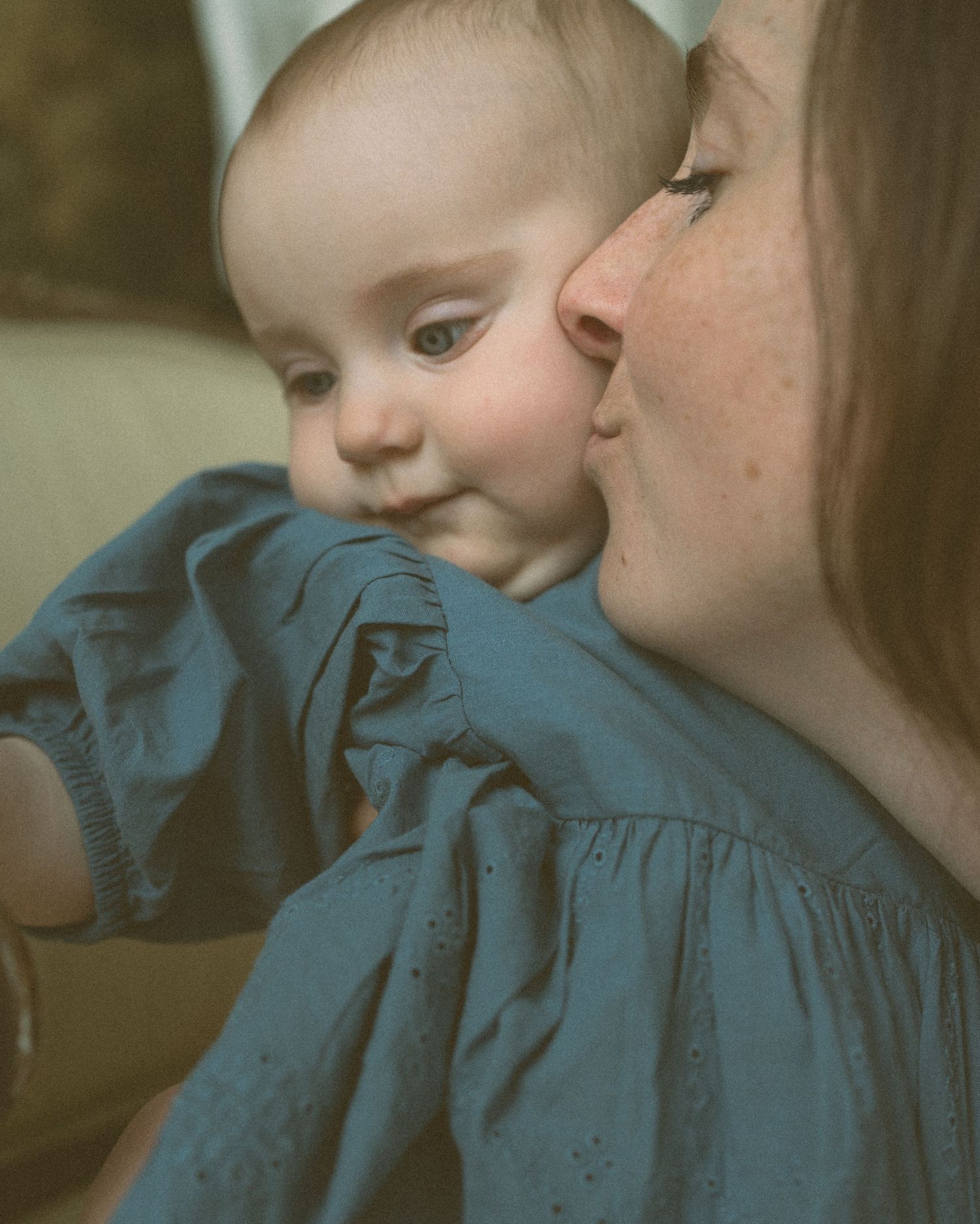A mother tenderly kisses her smiling baby dressed in a blue eyelet dress, captured in a warm, intimate portrait.
