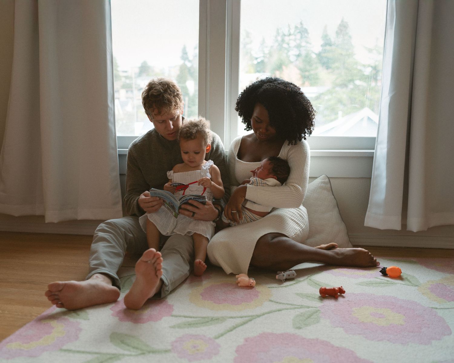 Family relaxing together on a pastel rug by large windows in a cozy living room.