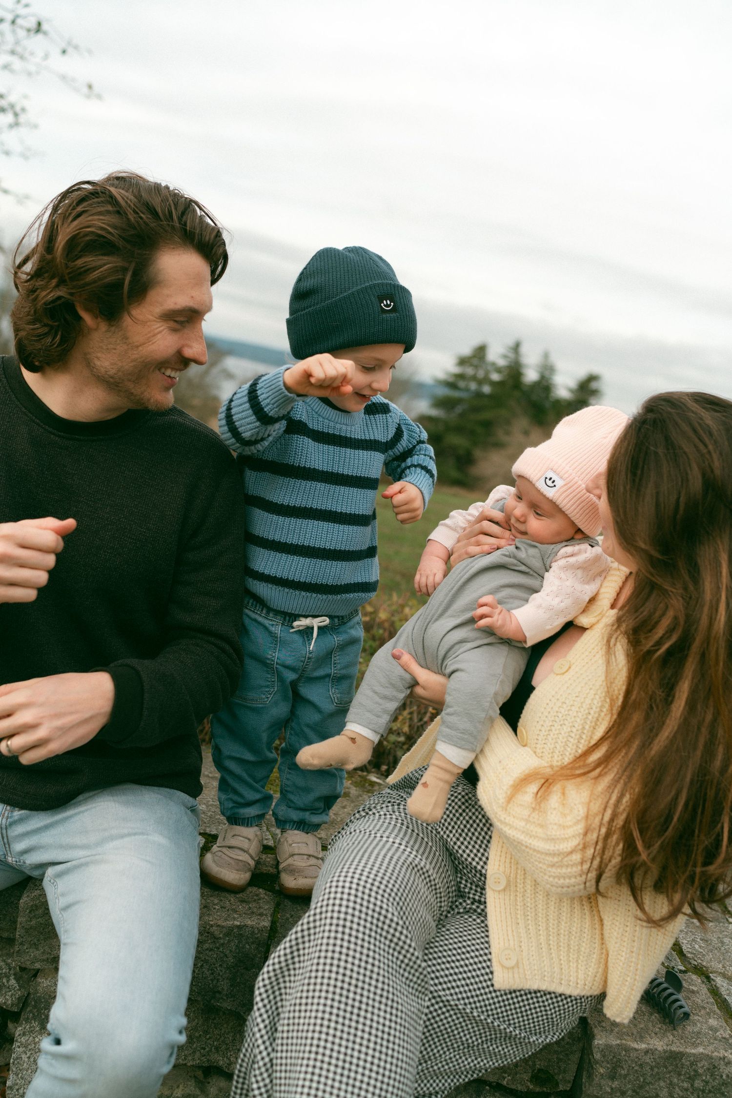 A family enjoys a playful moment together outdoors on a cloudy day, wearing casual sweaters and winter attire.