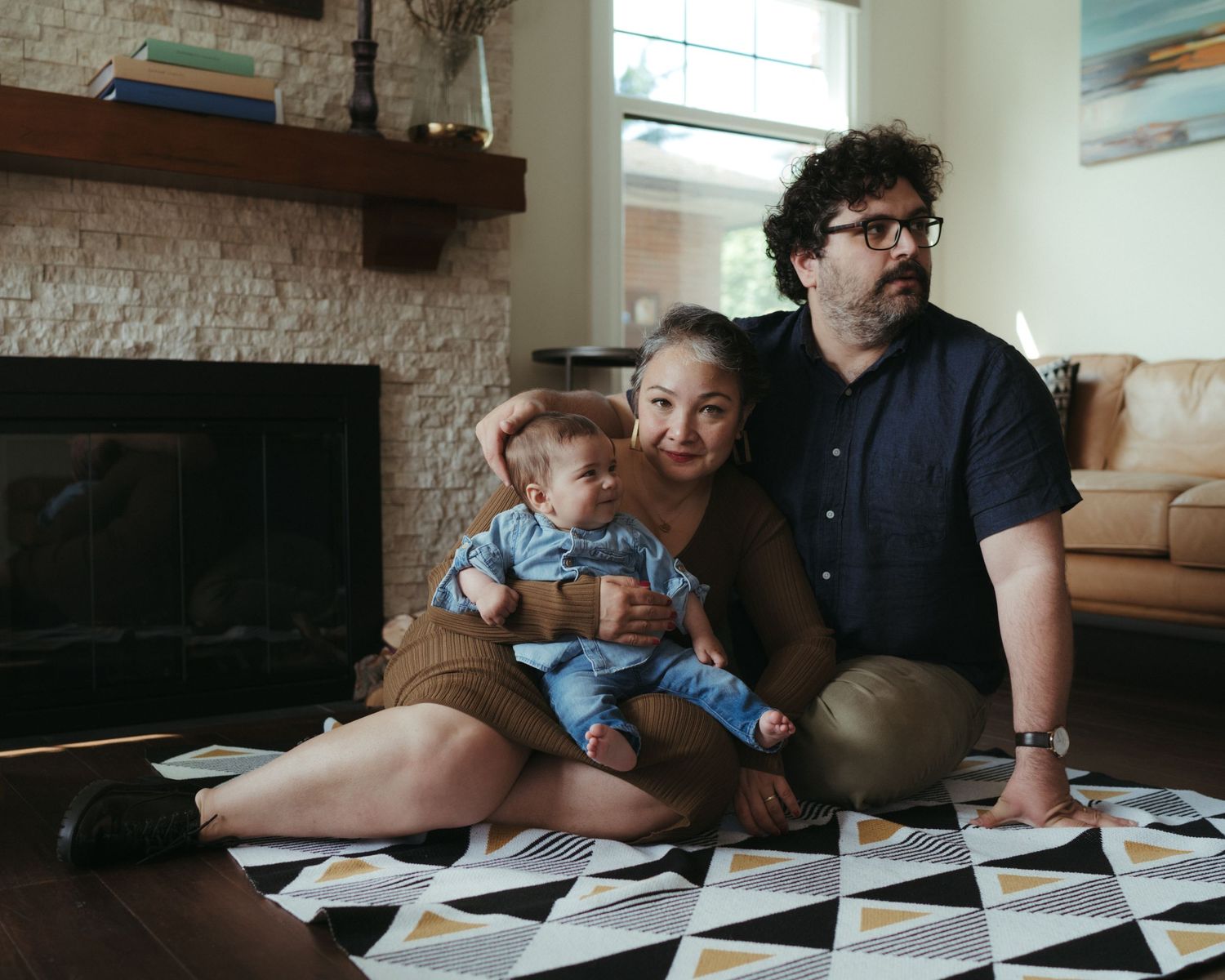 Family sits together on geometric patterned rug by brick fireplace in bright living room.