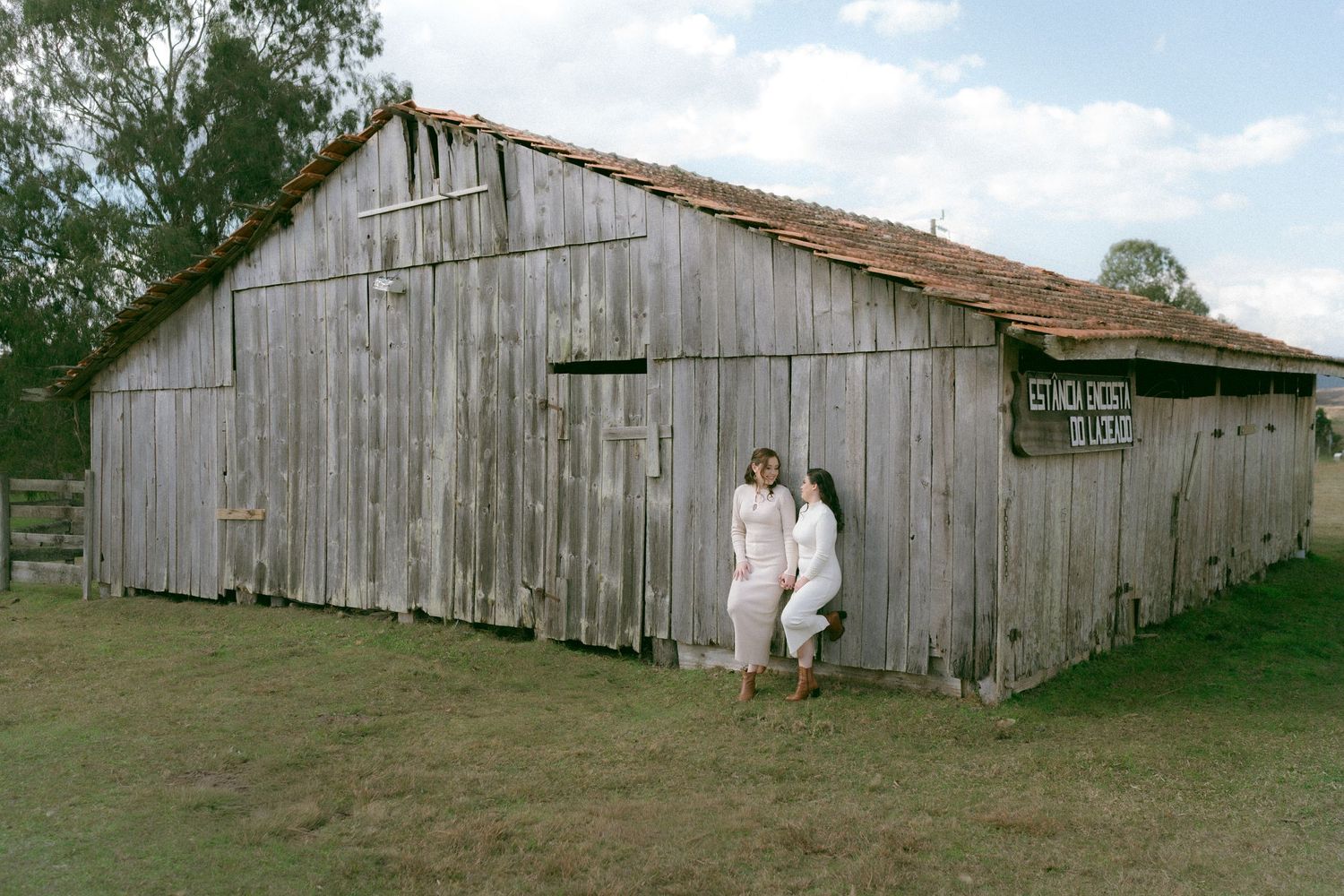Two women in white dresses pose outside a rustic weathered wooden barn on a green grass field.