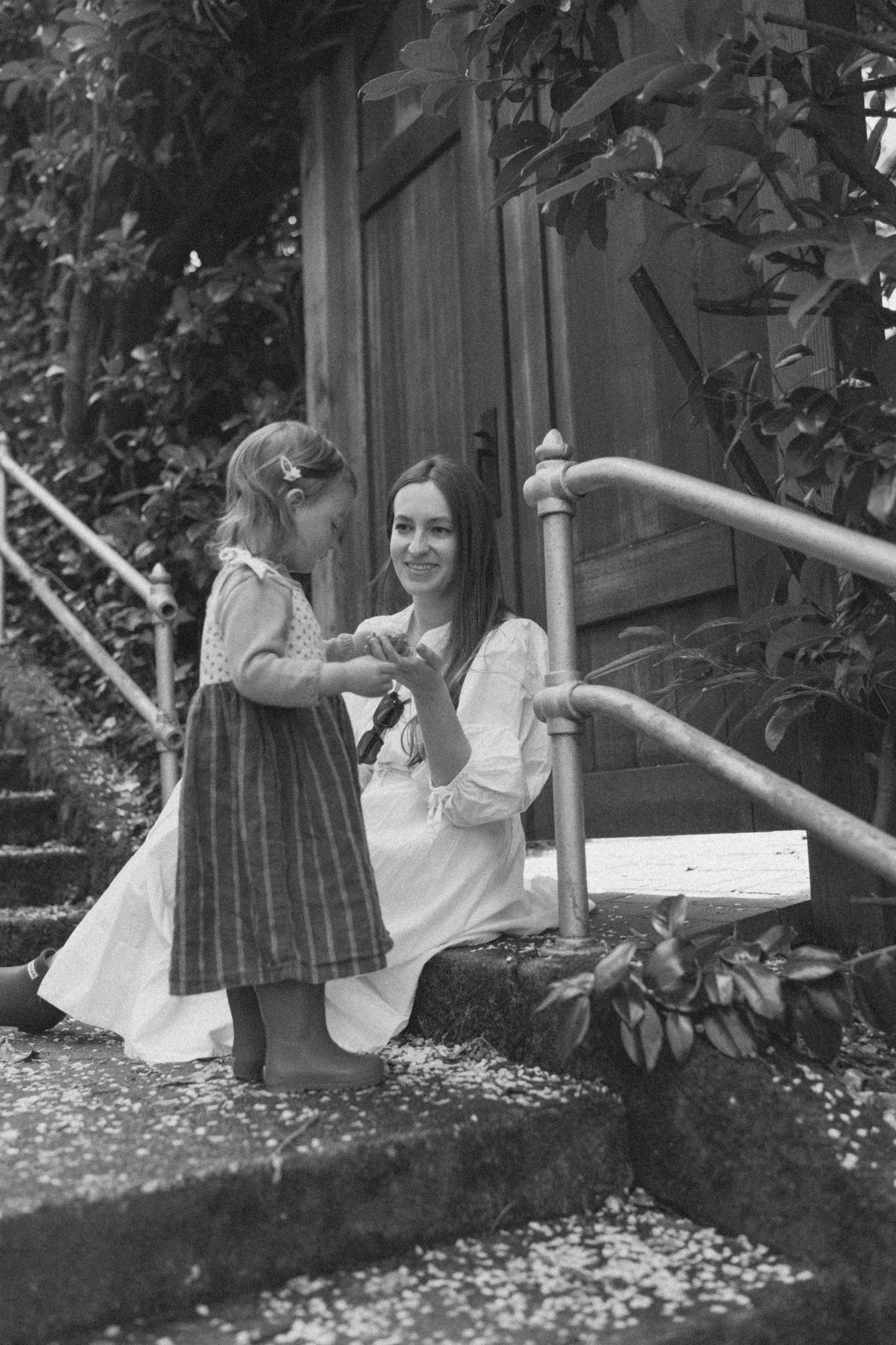 Black and white photo of a mother kneeling beside her young daughter on garden steps near a wooden door.