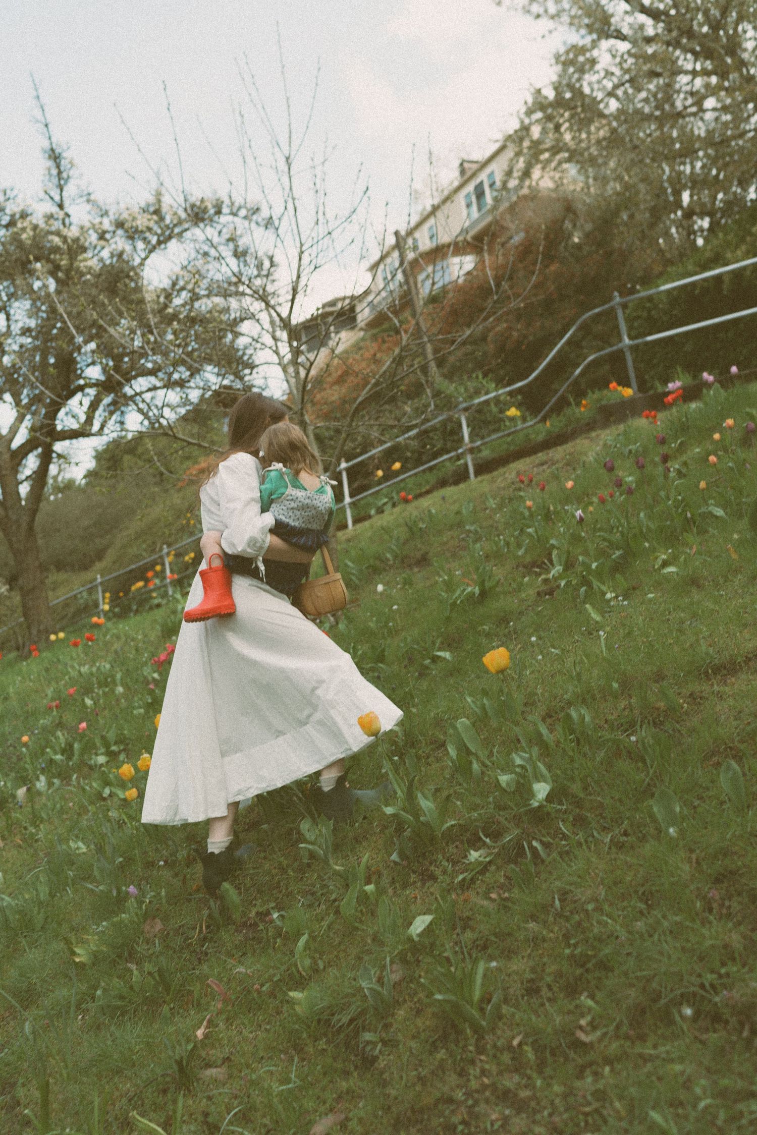 A woman in a white dress carries a child up a grassy hillside with colorful tulips and a house in the background.
