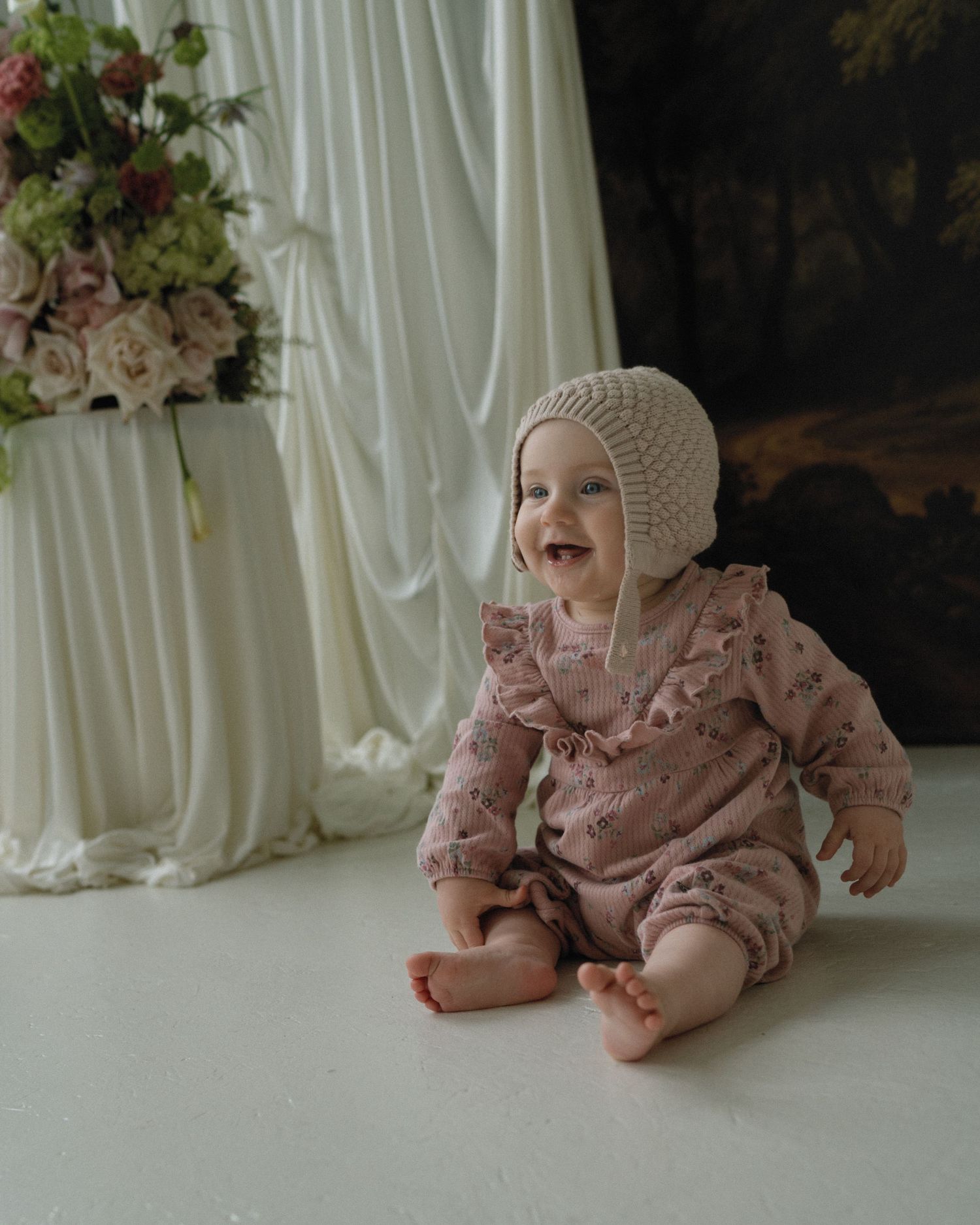 Smiling baby in floral romper and knit bonnet sits on white floor beside draped fabric and pink flower arrangement.