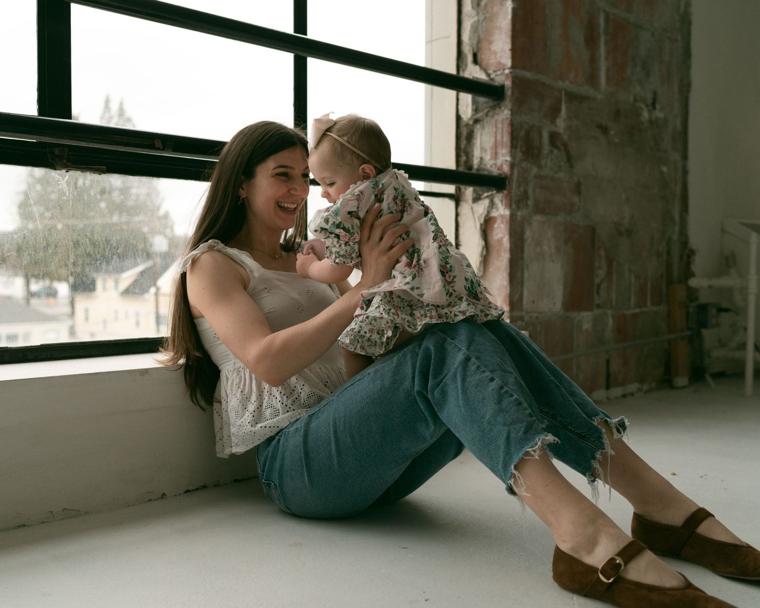 A young mother sitting on the floor by a window, smiling and holding her baby girl in a floral outfit.