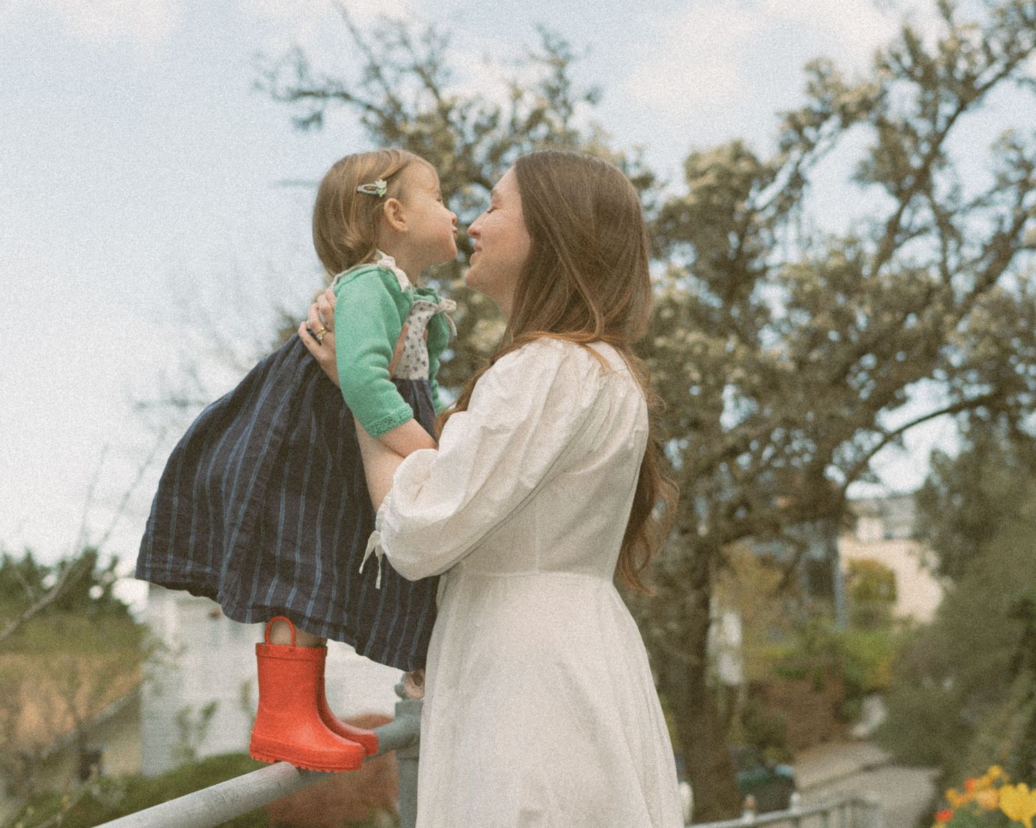 A young woman in a white dress holds a toddler in a plaid skirt and red boots outdoors.