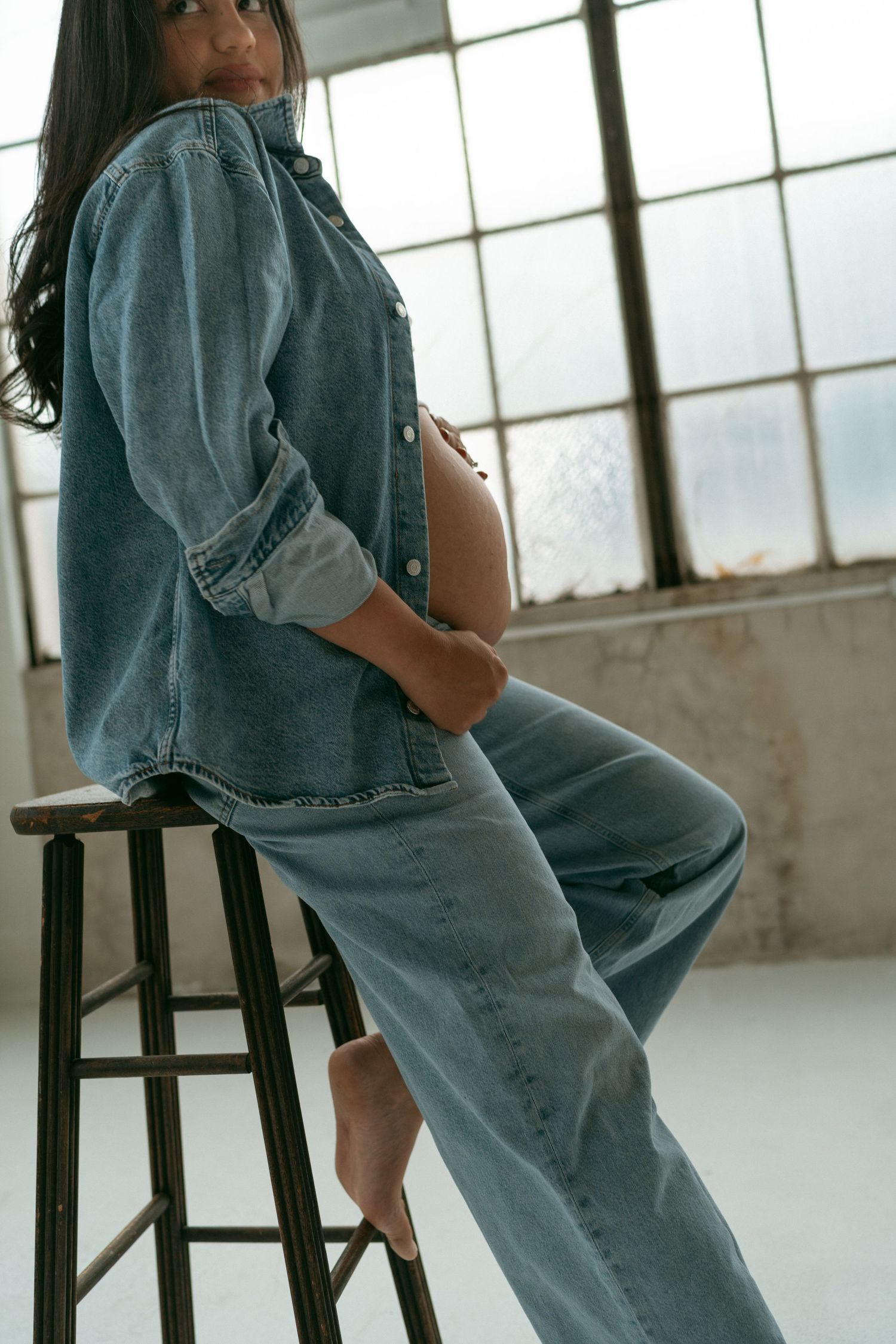 Pregnant woman in denim outfit sits on stool near window in artistic maternity photoshoot.