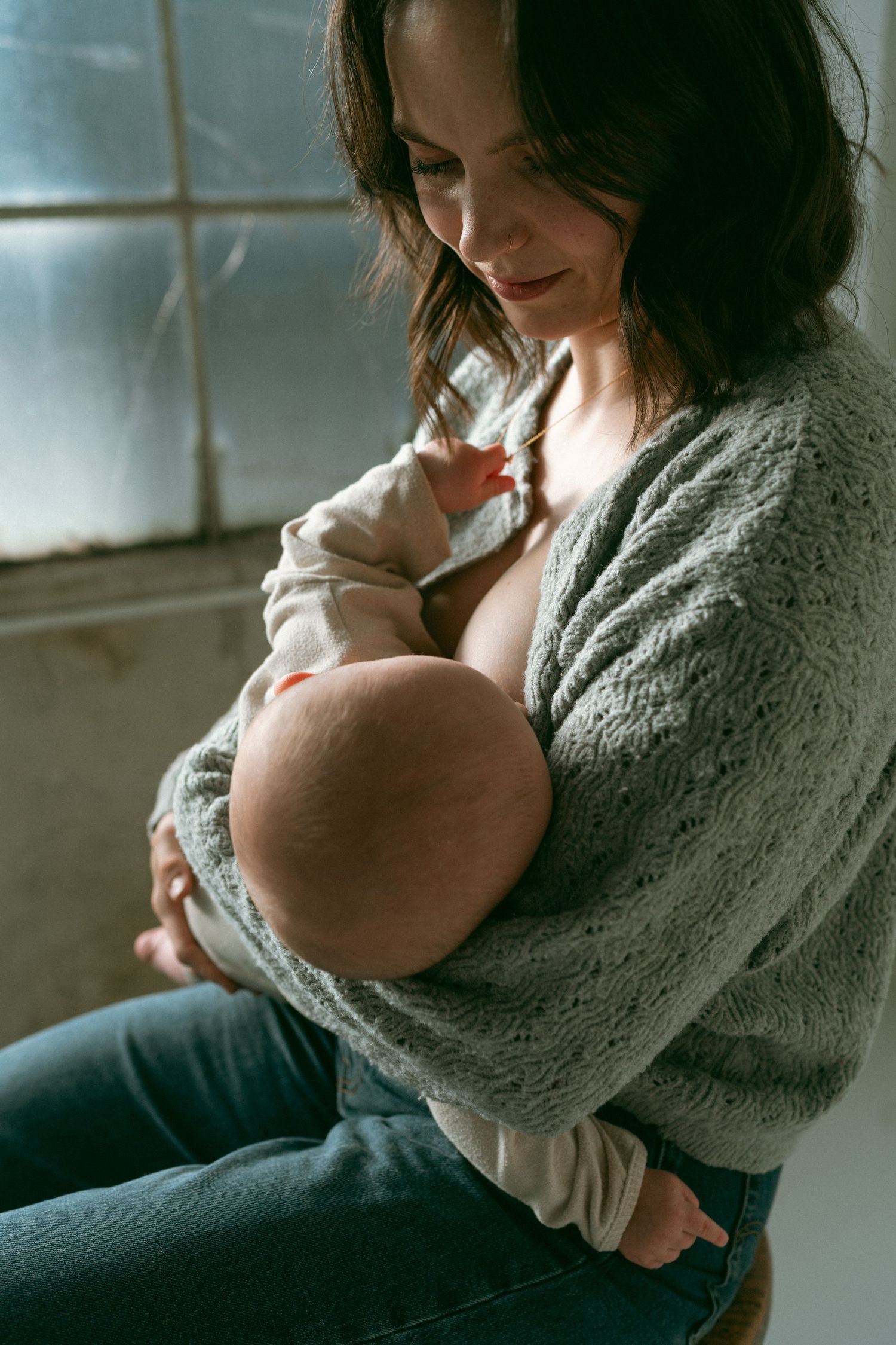 A mother in a gray sweater holds and nurses her newborn baby while sitting near a window in soft natural light.