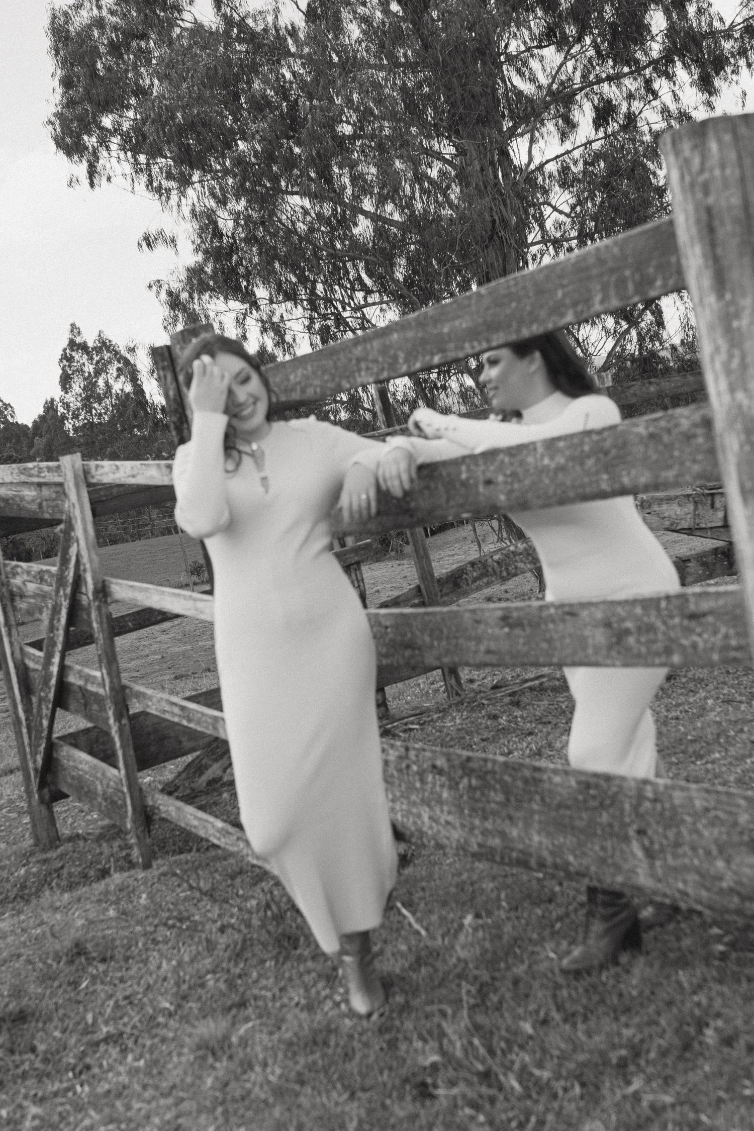 Two women in white dresses pose by a rustic wooden fence outdoors in a black and white photo.