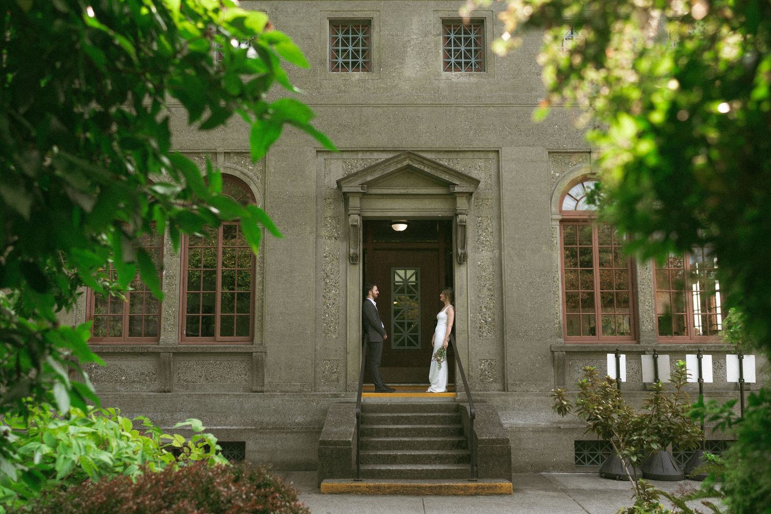 A grand stone building entrance framed by green foliage with a glimpse of a wedding dress through the doorway.