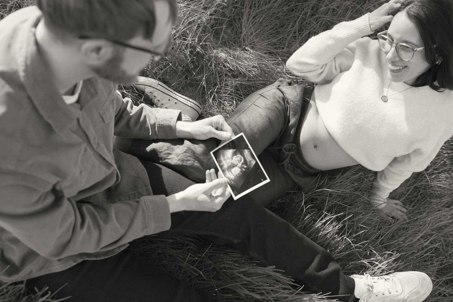 A couple looks at ultrasound photos while sitting in grass.