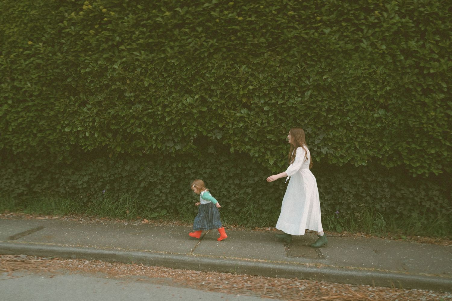 A woman in a white dress walks with a toddler in red boots along a sidewalk beside a lush green hedge.
