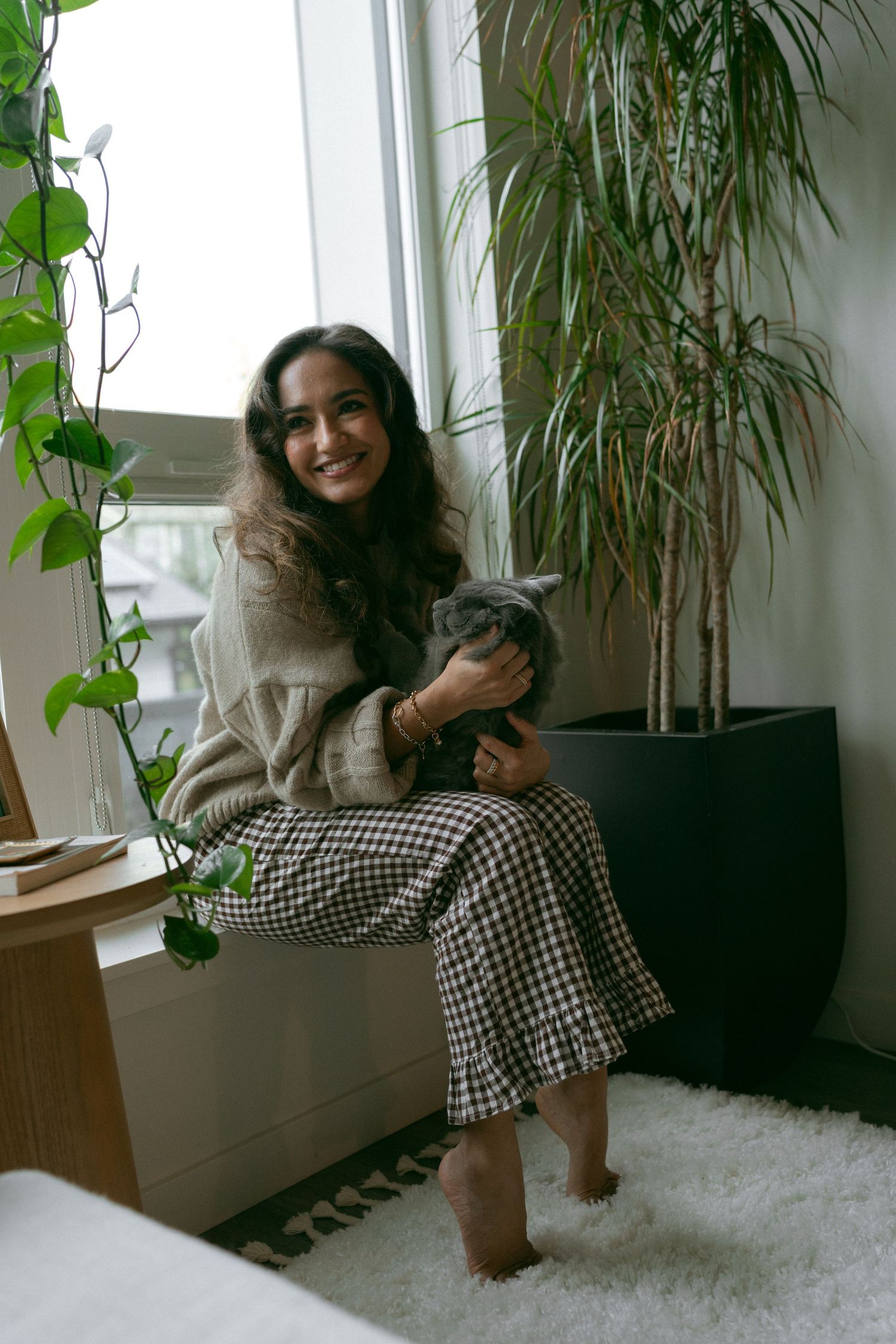Person and grey cat lounging by window with indoor plants and white carpet.