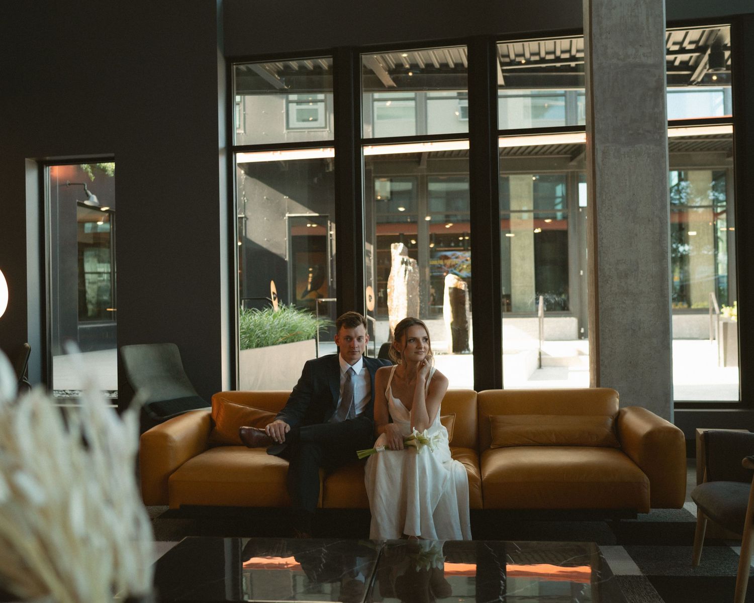 A bride and groom sit together on a tan leather sofa in a modern lobby with floor-to-ceiling windows and a reflective glass table.