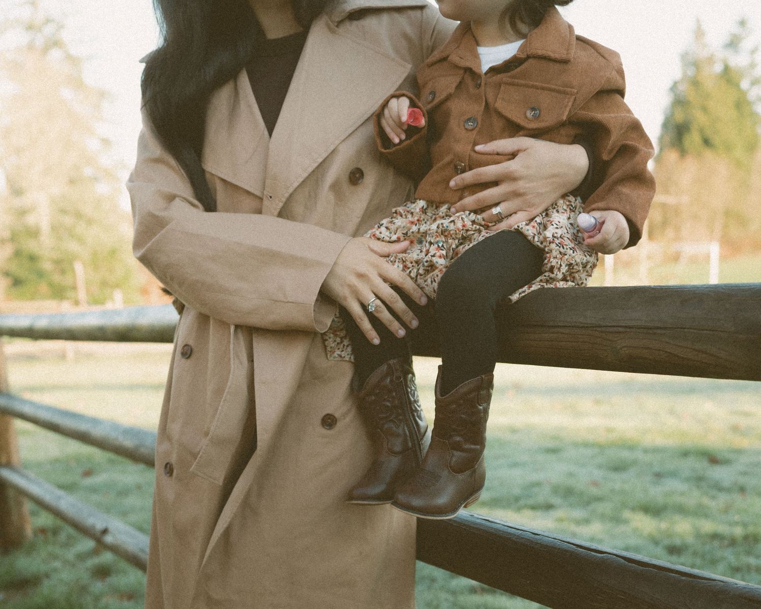 A person wearing a beige coat holds a small dog while sitting on a wooden bench in an outdoor setting.