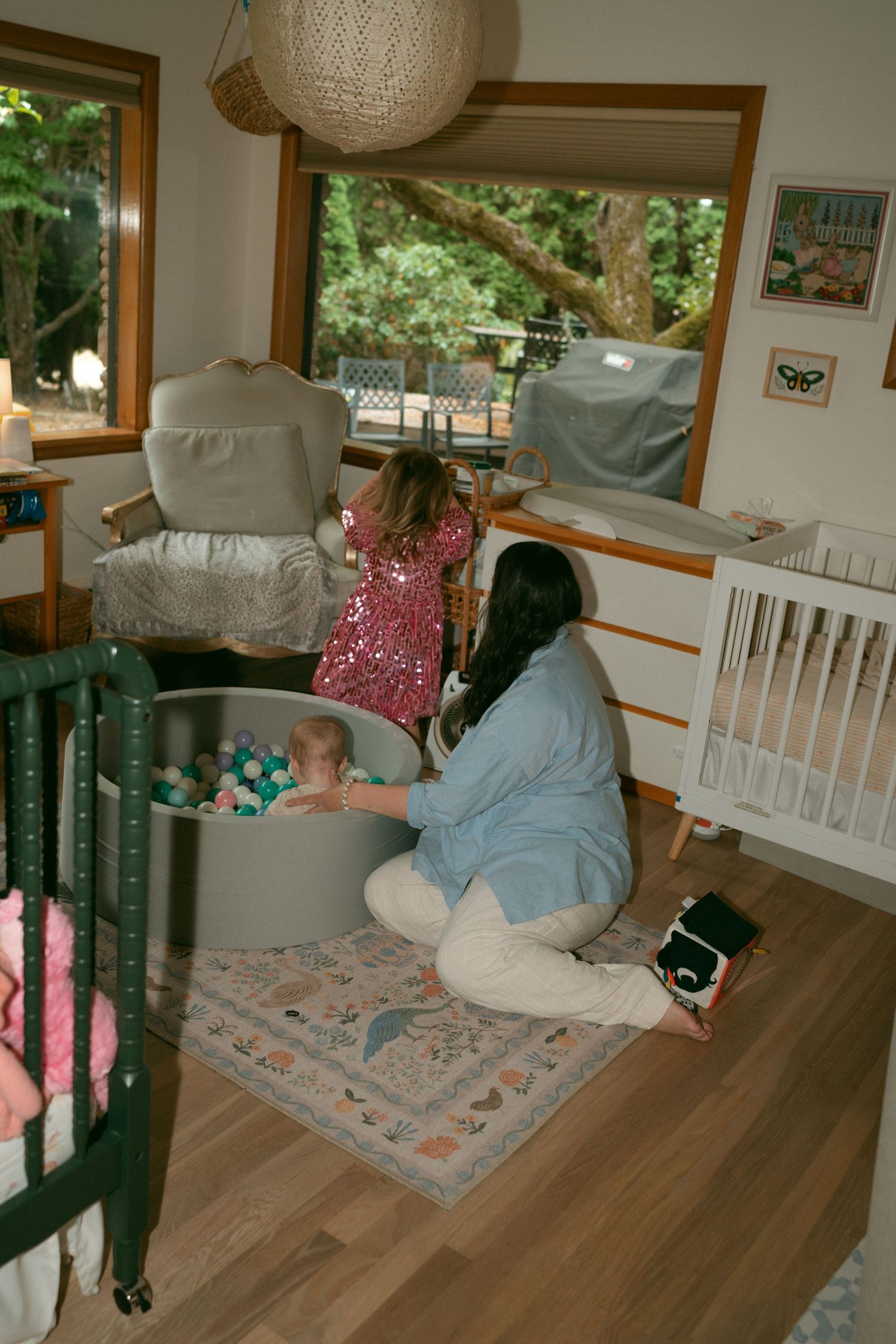 A woman sits on a patterned rug beside a baby in a ball pit while a girl in a sparkly pink dress looks out the window in a bright nursery.