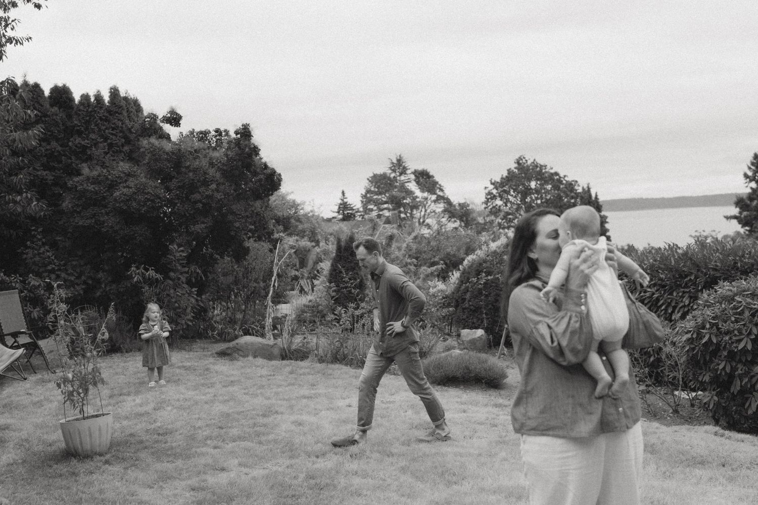 Black and white photo of a family playing on a backyard lawn by the water, with a mother holding a baby while a man walks nearby.