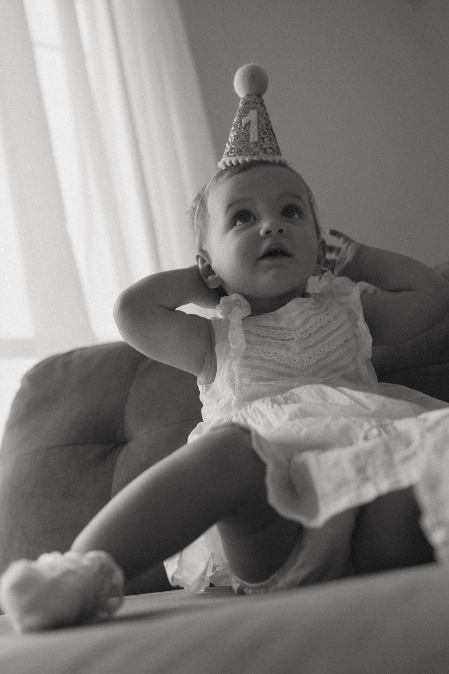 Black and white photo of a baby girl in a white dress and party hat sitting on a couch looking up.