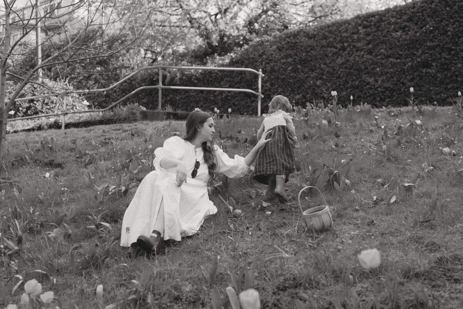 Black and white photo of a woman in white dress helping a young girl pick flowers in a garden.